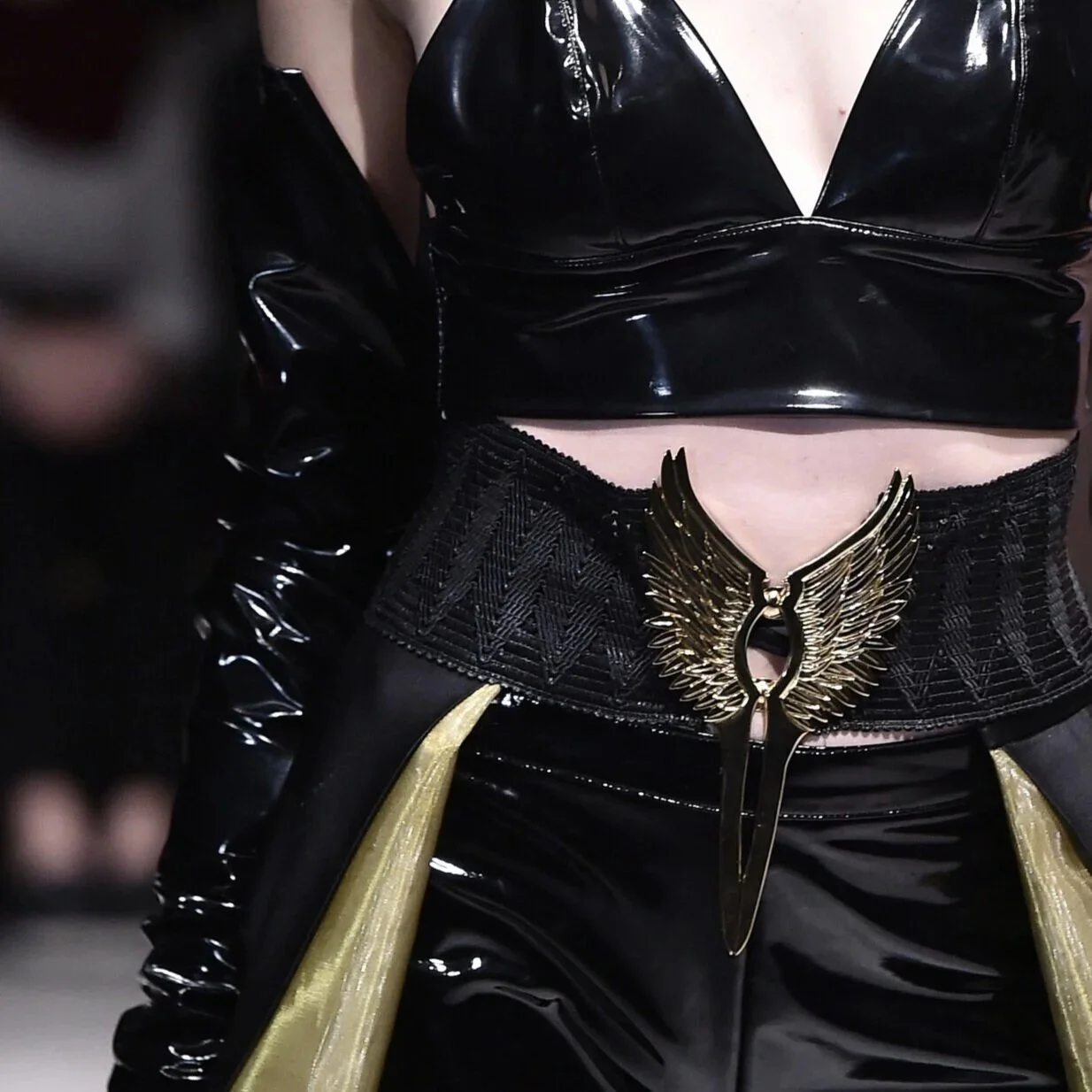 Close-up of a person wearing a black, shiny, vinyl crop top and a wide belt with gold angel wing design, in a futuristic or fantasy style fashion.