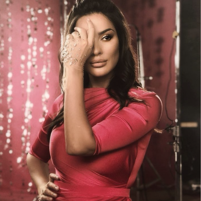 A woman with dark hair, wearing a red dress, is posing with one hand on her hip and the other hand covering part of her face, in front of a pink background with hanging shiny decorations.