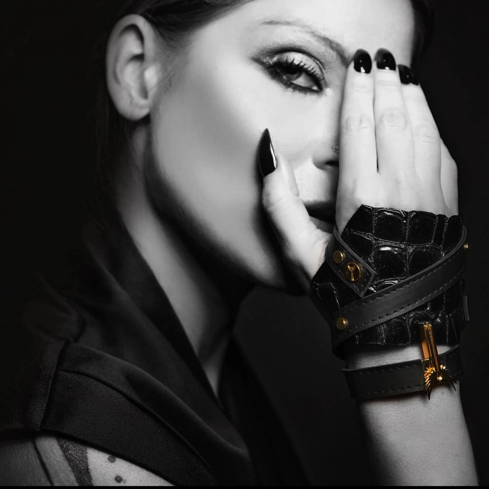 A black and white close-up of a woman with makeup and long nails, covering one eye with her hand. She is wearing leather bracelets with gold accents.