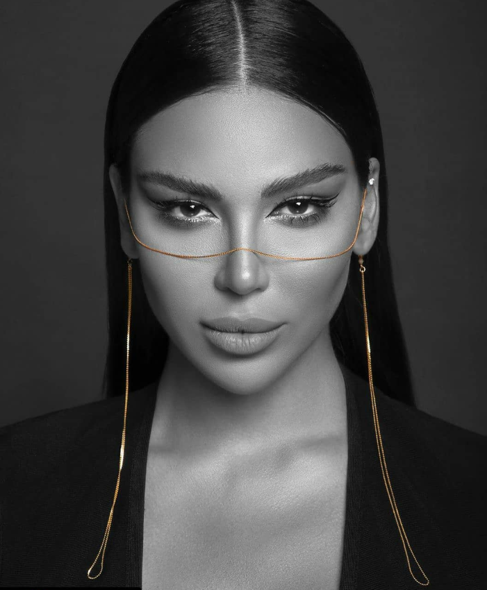 Black and white portrait of a woman with long, straight hair, wearing gold face chains and earrings, looking directly at the camera.