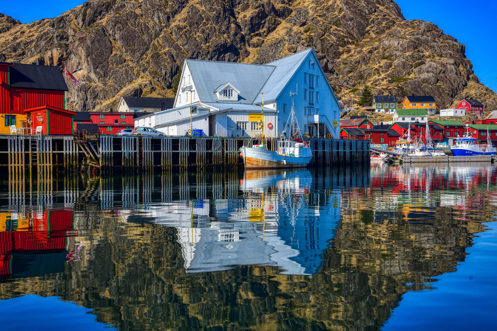Compose a stunning image capturing the scenic route of Tromsø to Lofoten. Highlight the grand jagged peaks of Lofoten, with clear, turquoise waters below. Infuse the scene with dynamic, scattered clouds and soft sunlight, ideal for showcasing the dramatic landscape. The image should portray a charter boat on the move, with adventurous travelers onboard. Consider a slightly elevated perspective that reveals the vastness of the coastline and the allure of untouched nature. Ensure the textures on the boat and water are extremely clear and atmospheric.