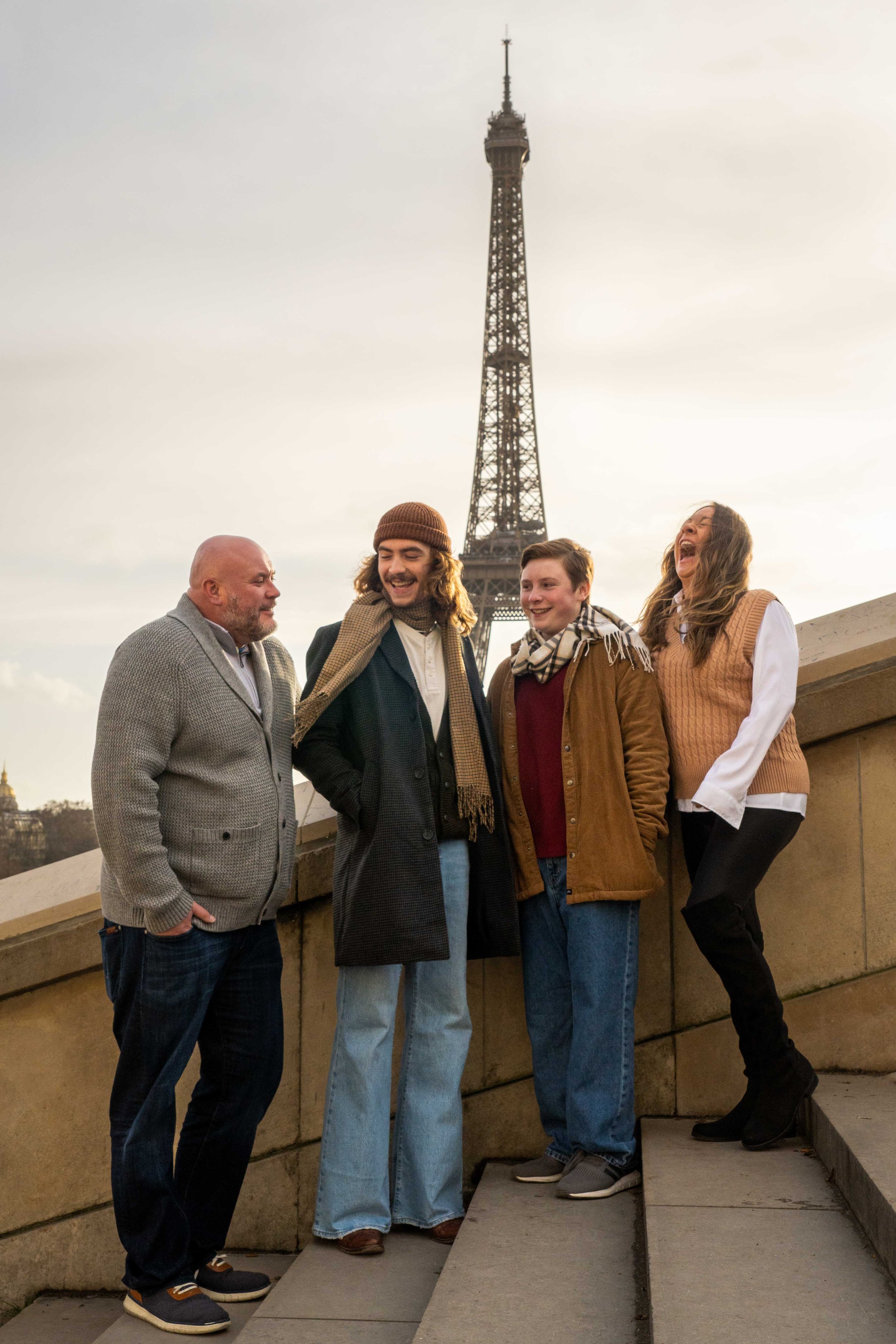 A group of four people standing on steps near the Eiffel Tower in Paris, France, laughing and enjoying each other's company during what appears to be a sunny day.
