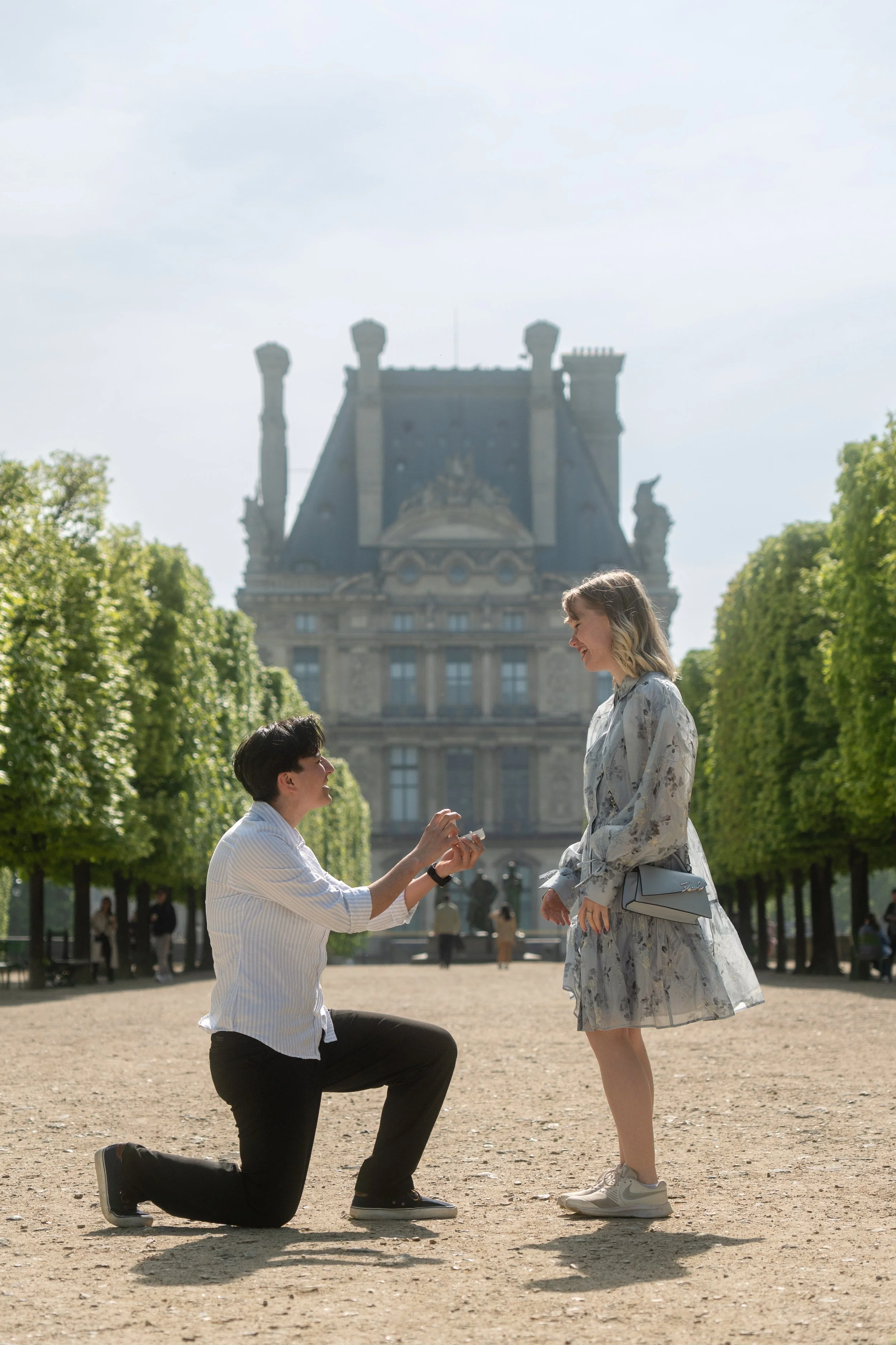 A man proposing marriage to a woman in a park with a historic building in the background.