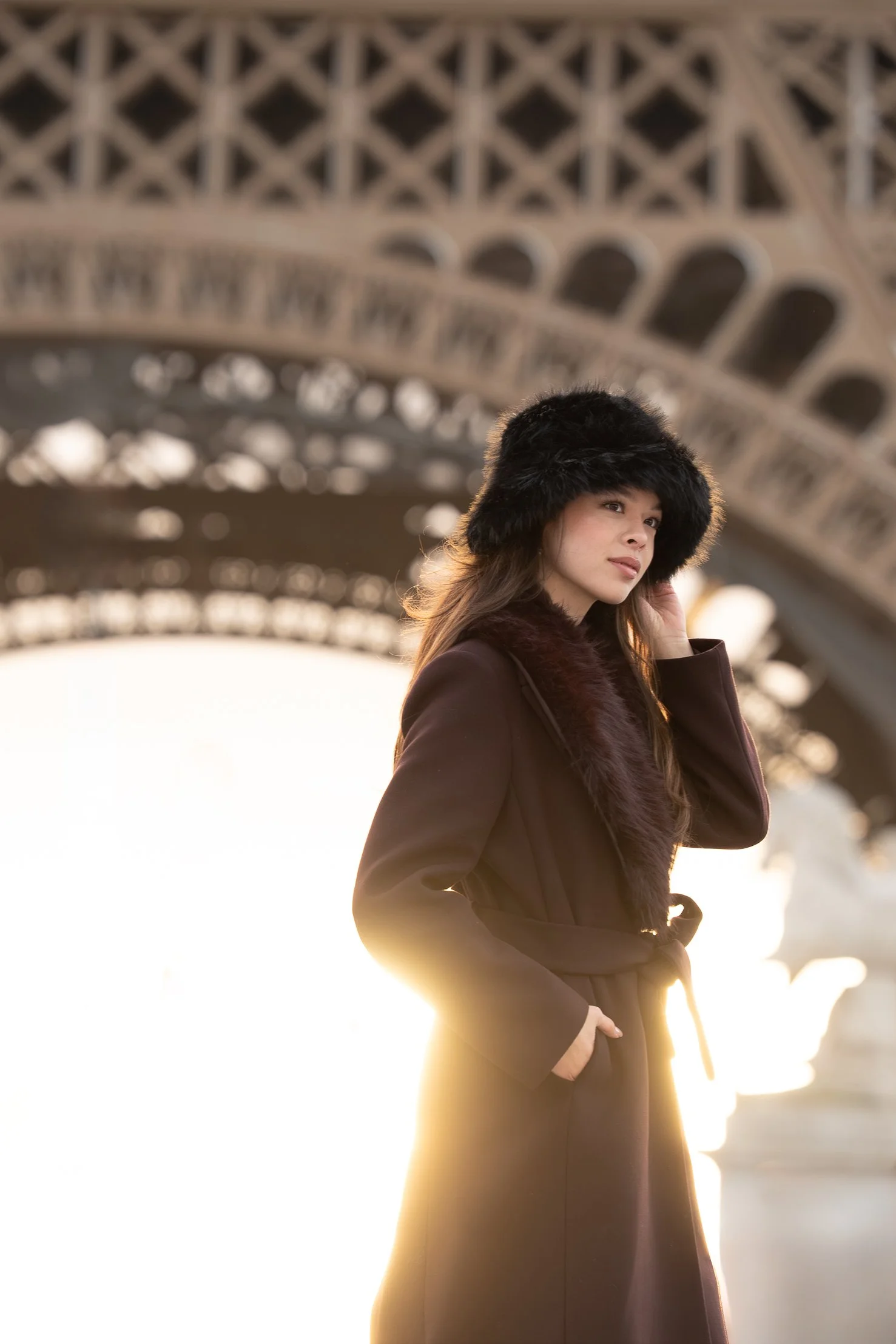 A woman in a dark coat with a fur collar and hat standing near the Eiffel Tower in Paris, France during sunset.