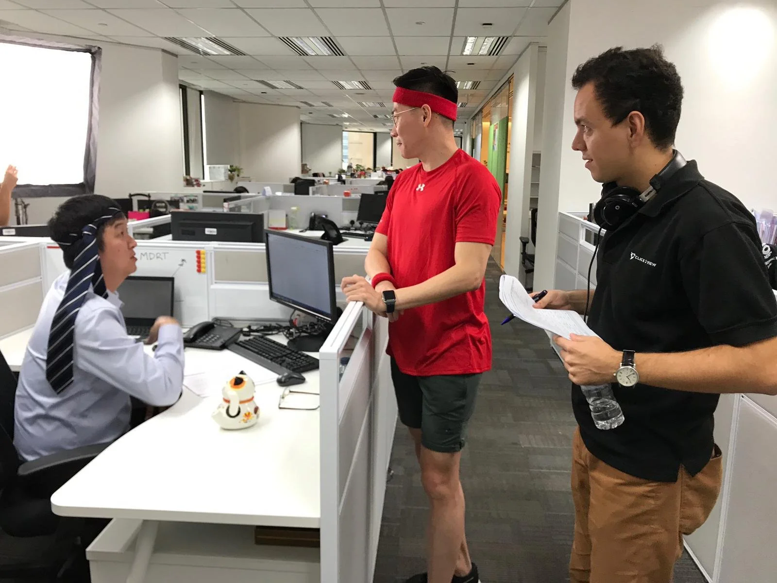 Office environment with a woman in a white shirt and tie sitting at a desk, talking to two men standing in front of her. The man in the red shirt and shorts is wearing a red headband and smartwatch. The other man is holding a water bottle, paper, and