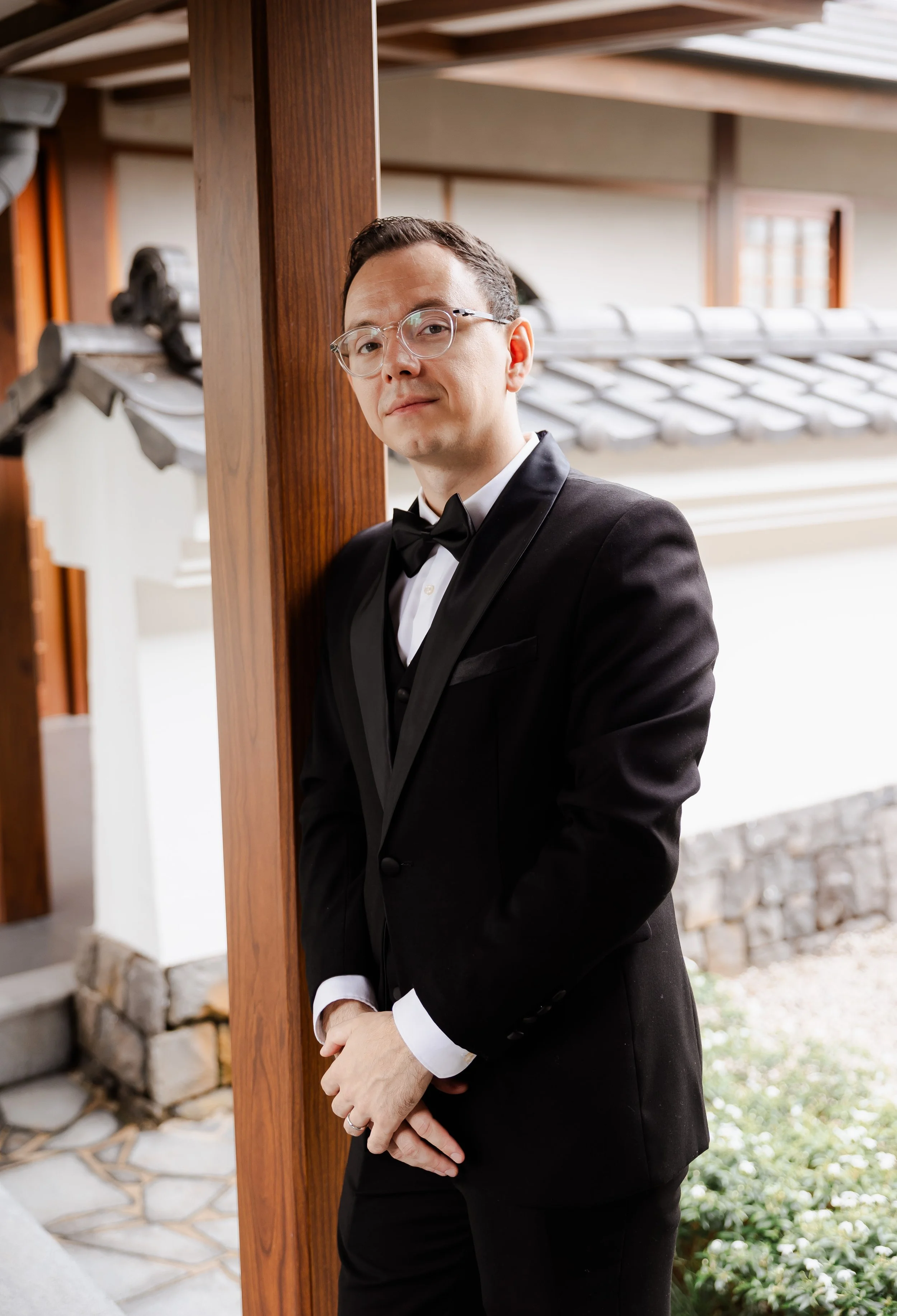 A man in a black tuxedo with a bow tie stands outdoors, leaning against a wooden post with a traditional Japanese roof in the background.