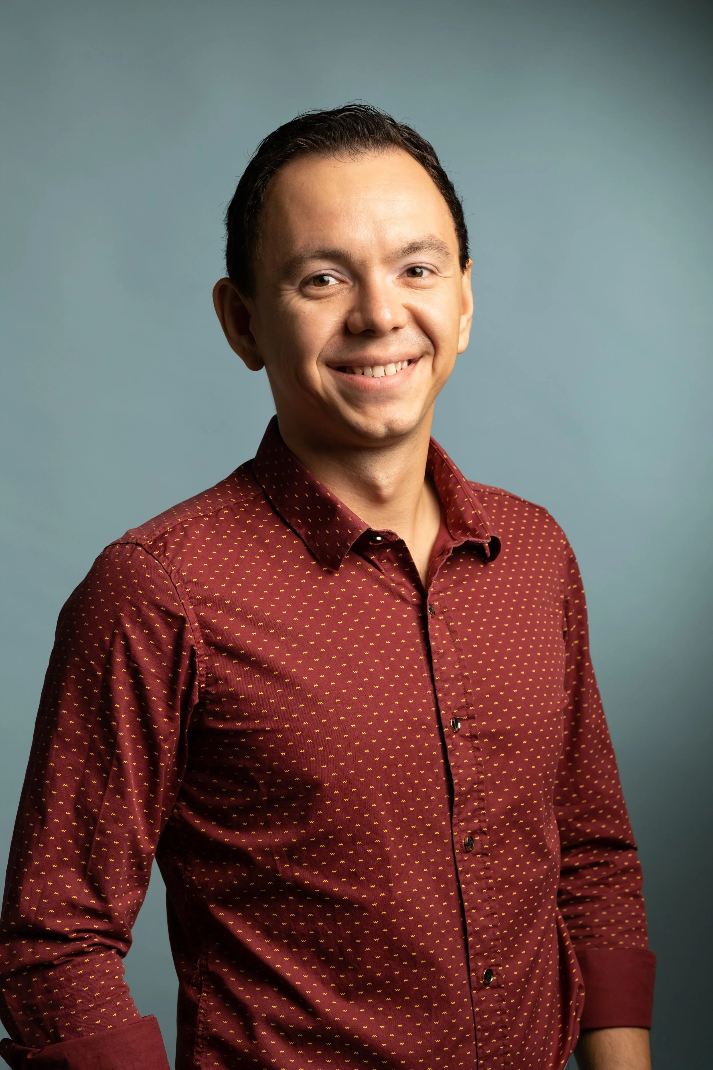 Portrait of a man with short dark hair, smiling, wearing a burgundy shirt with small yellow patterns, against a plain teal background.