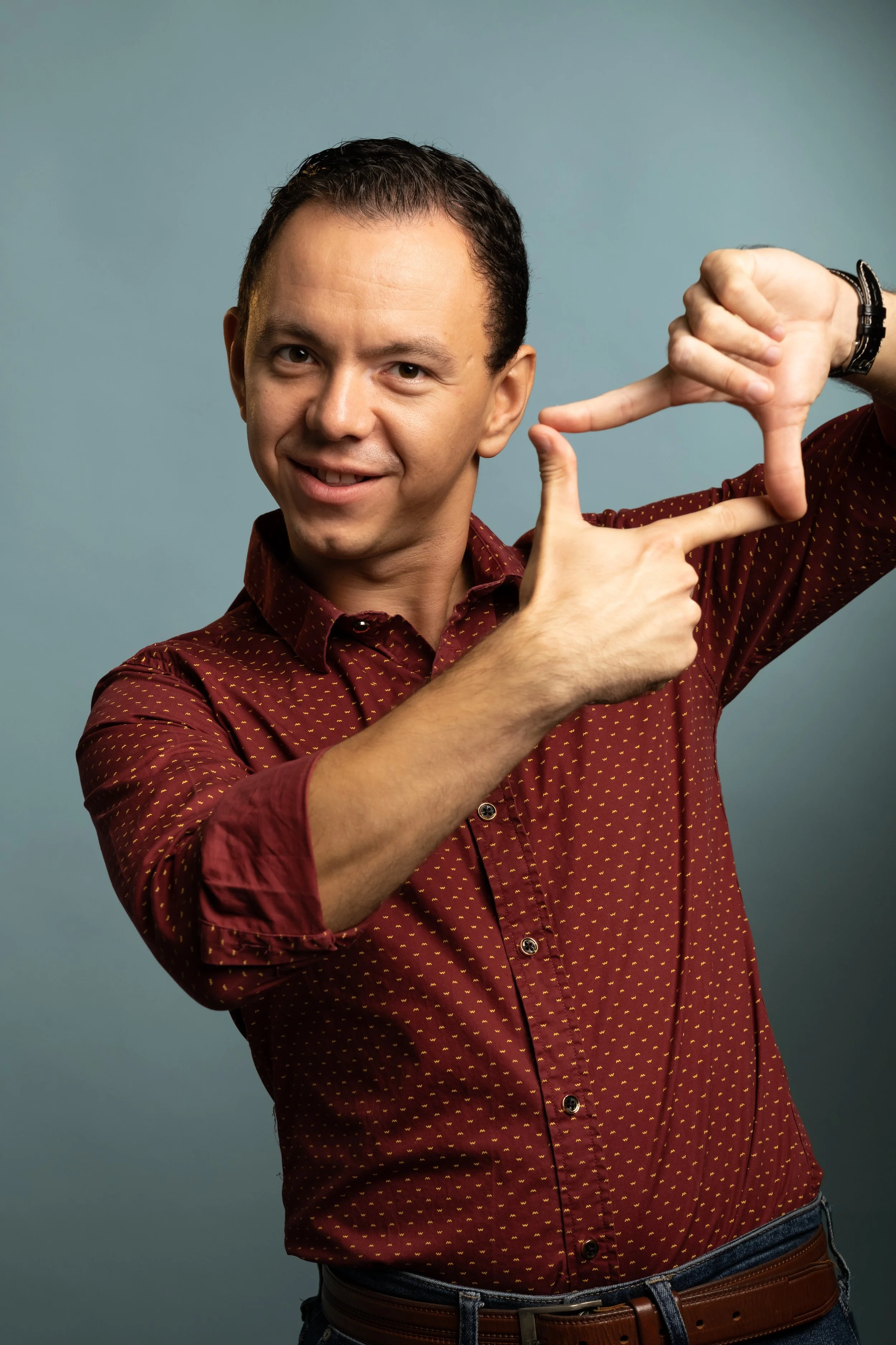 A man smiling and making a framing gesture with his hands, against a plain background.