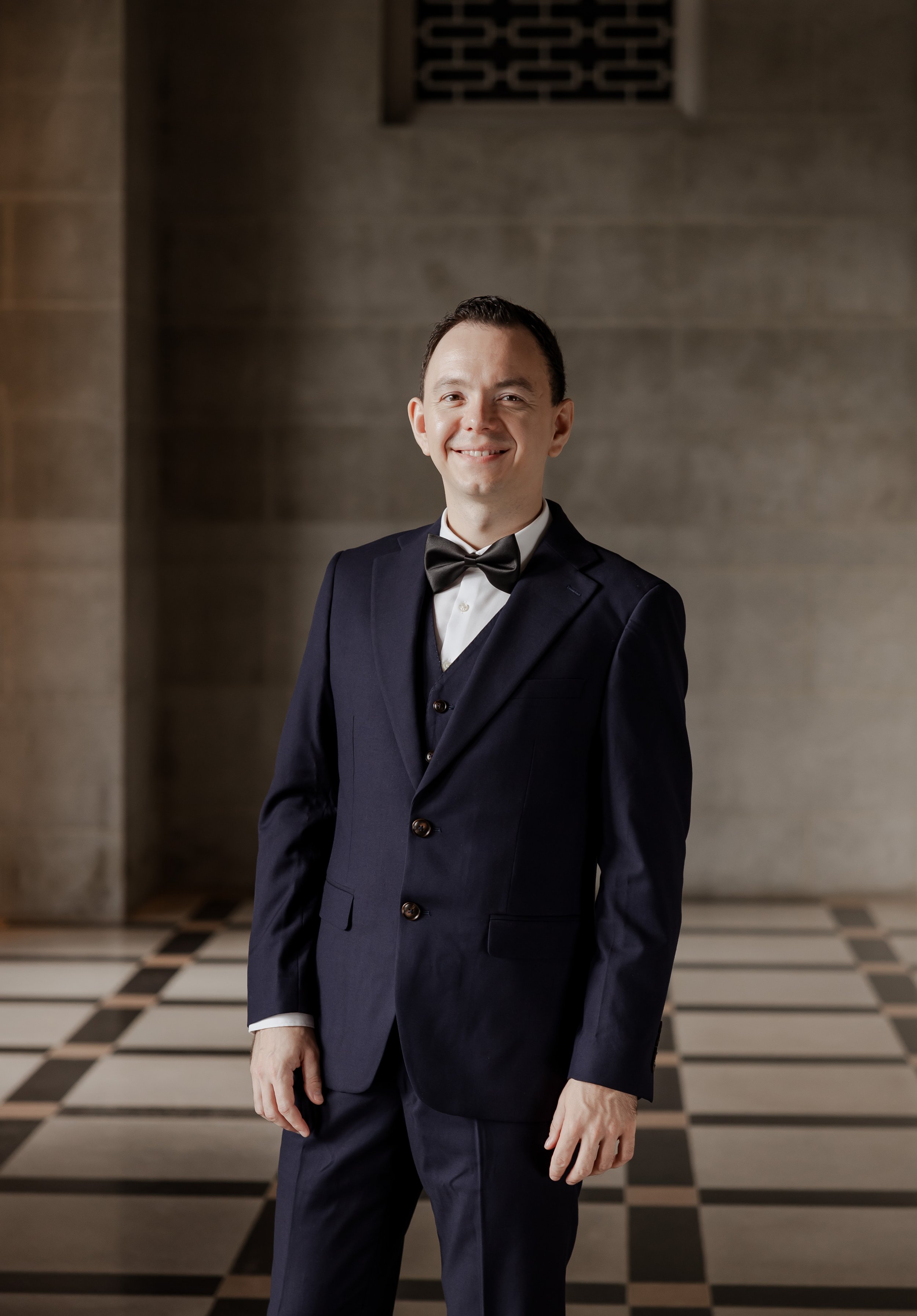 A man in a navy blue tuxedo with a black bow tie, standing indoors in front of a stone wall with a patterned floor, smiling at the camera.
