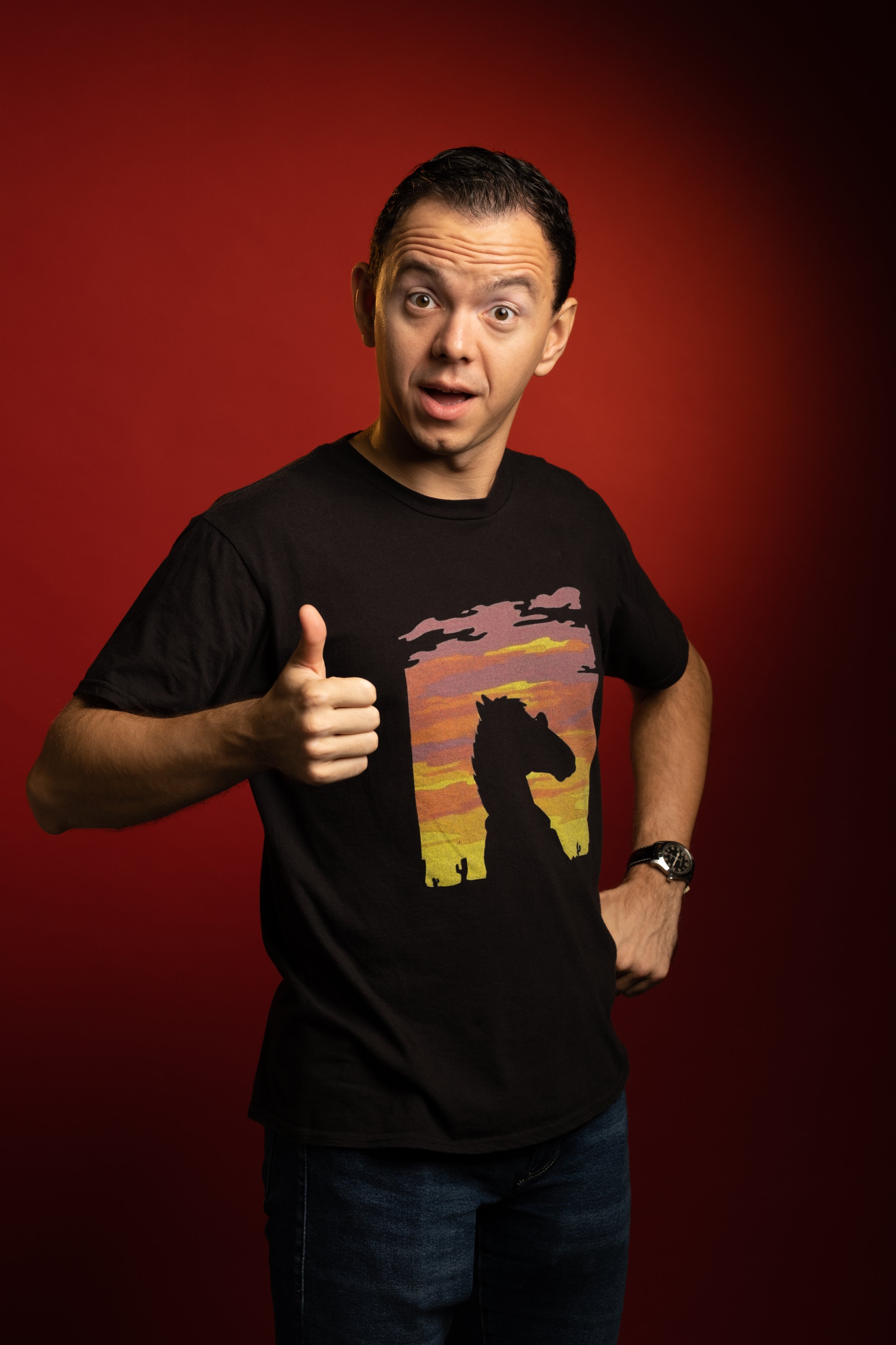 Man in black t-shirt with a desert sunset and cacti on it, giving a thumbs up against a red background.