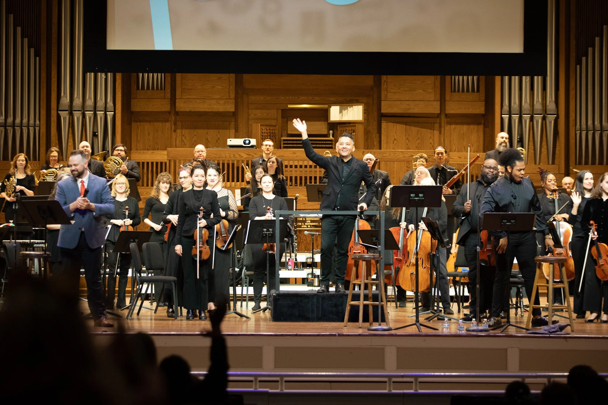 Music Director John Concklin stands onstage with musicians and waves to the Link Up audience