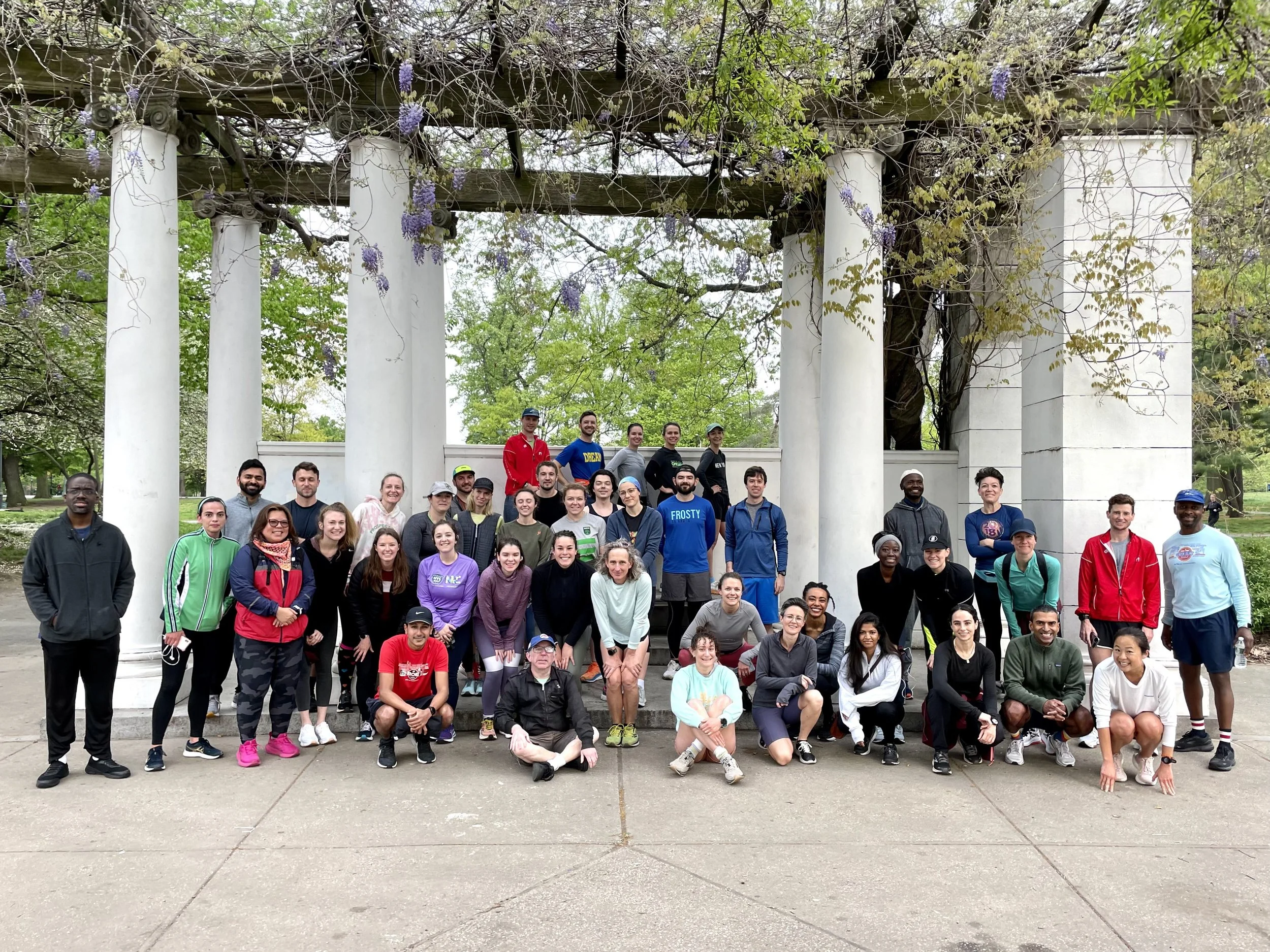 A group of about 45 runners sitting, squatting and standing underneath a pergola covered in vines and wisteria flowers. The runners in the very back are all posing with their hands on their hip. The runners are smiling.