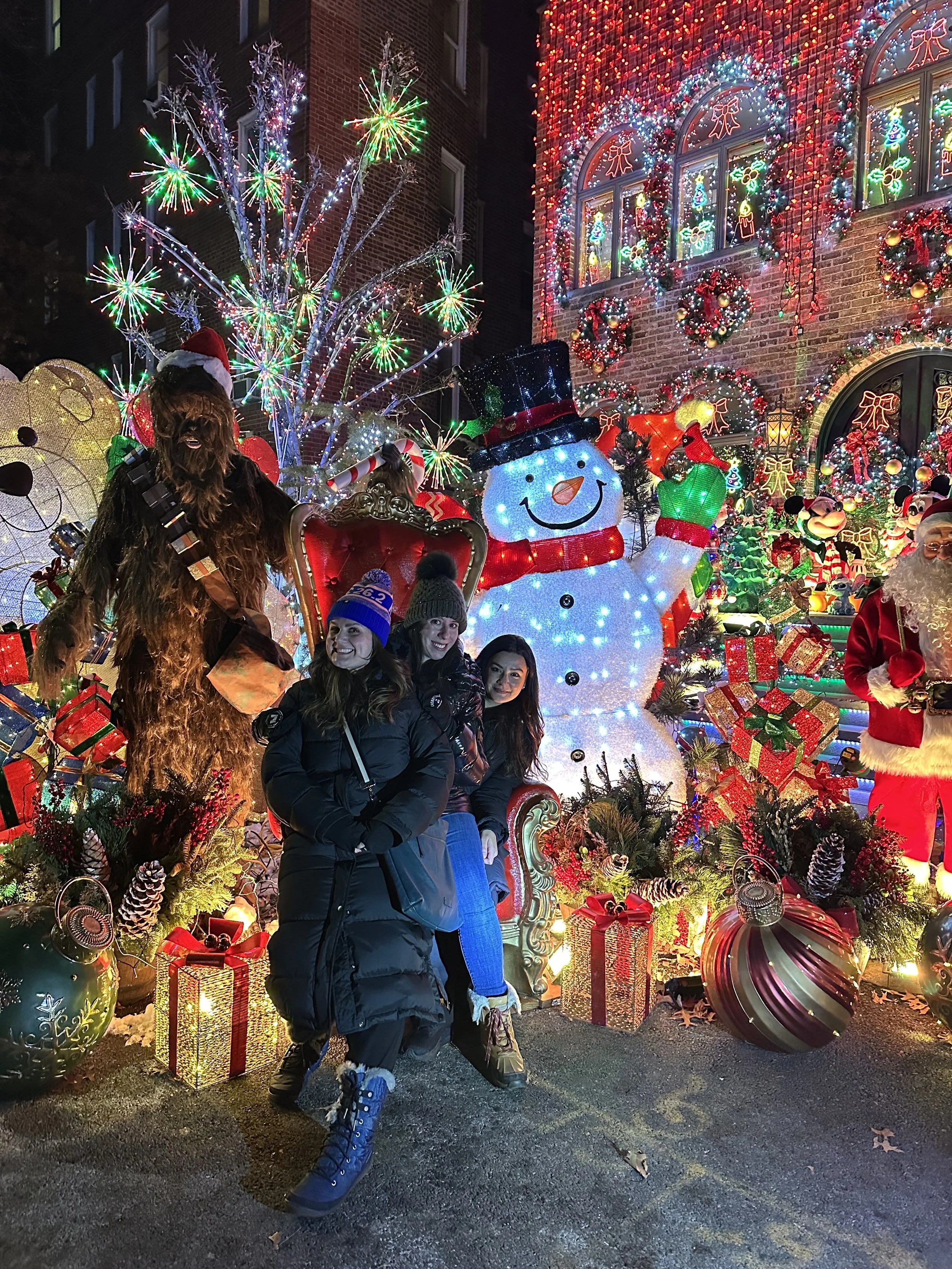 In front of a display of Christmas lights including a snowman, Santa, and Chewbacca, stand three smiling women, 2 of whom are wearing winter hats with pom-poms on top.