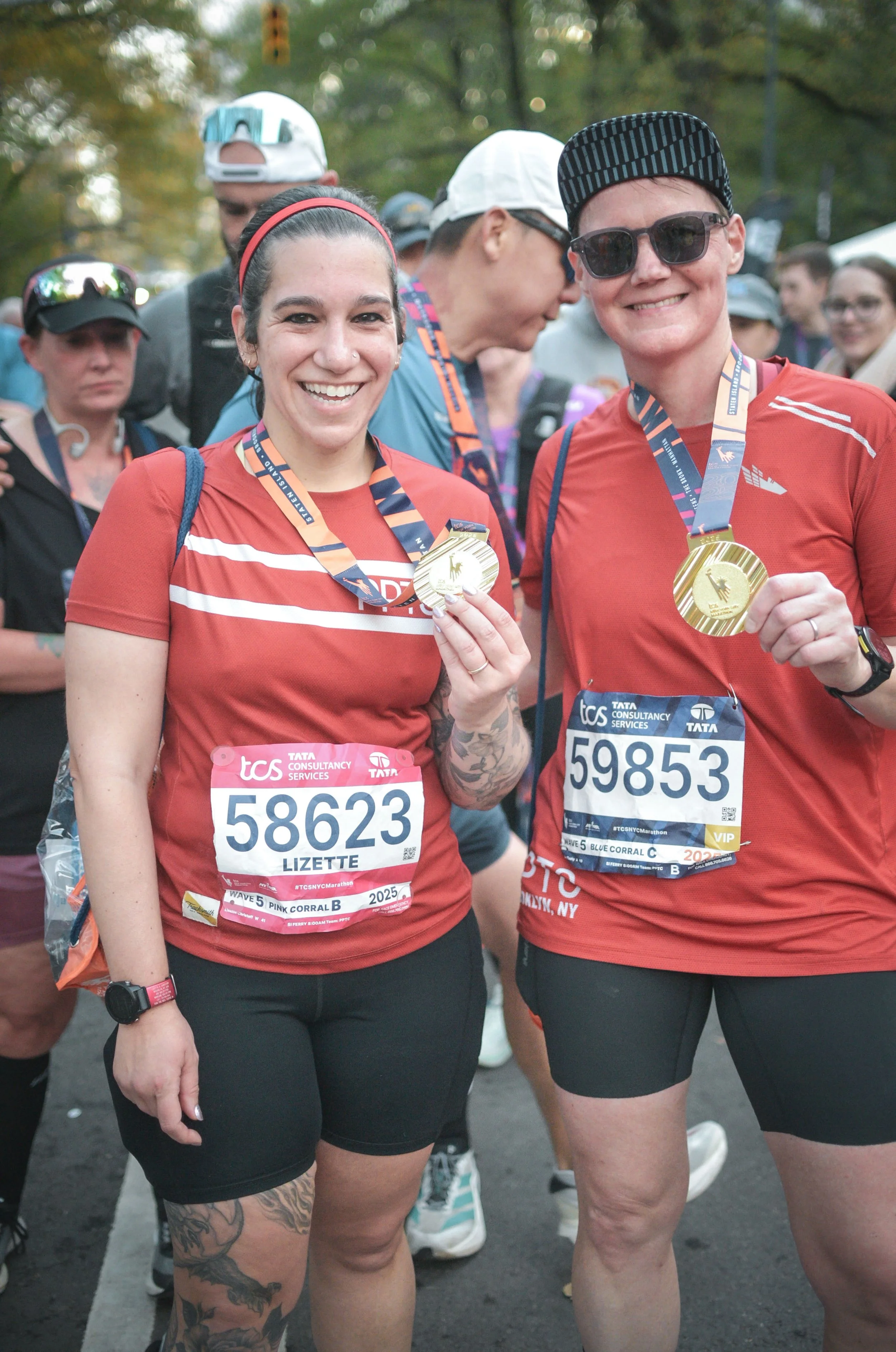 Two people in red running shirts hold up their medals and smile as other people mill about in the background.