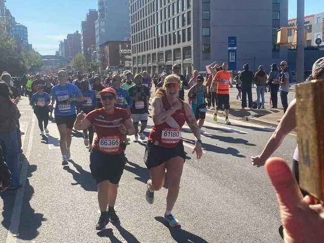 Two people run and smile in red race shirts as a crowd of people behind them are also running.