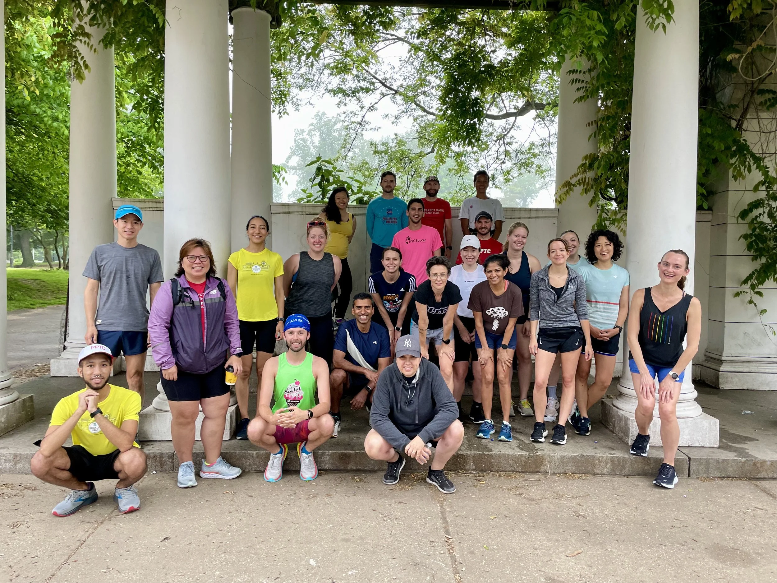 A group of 20-23 people in run clothes stand underneath a pergola covered in vines of verdant plants. They are all smiling