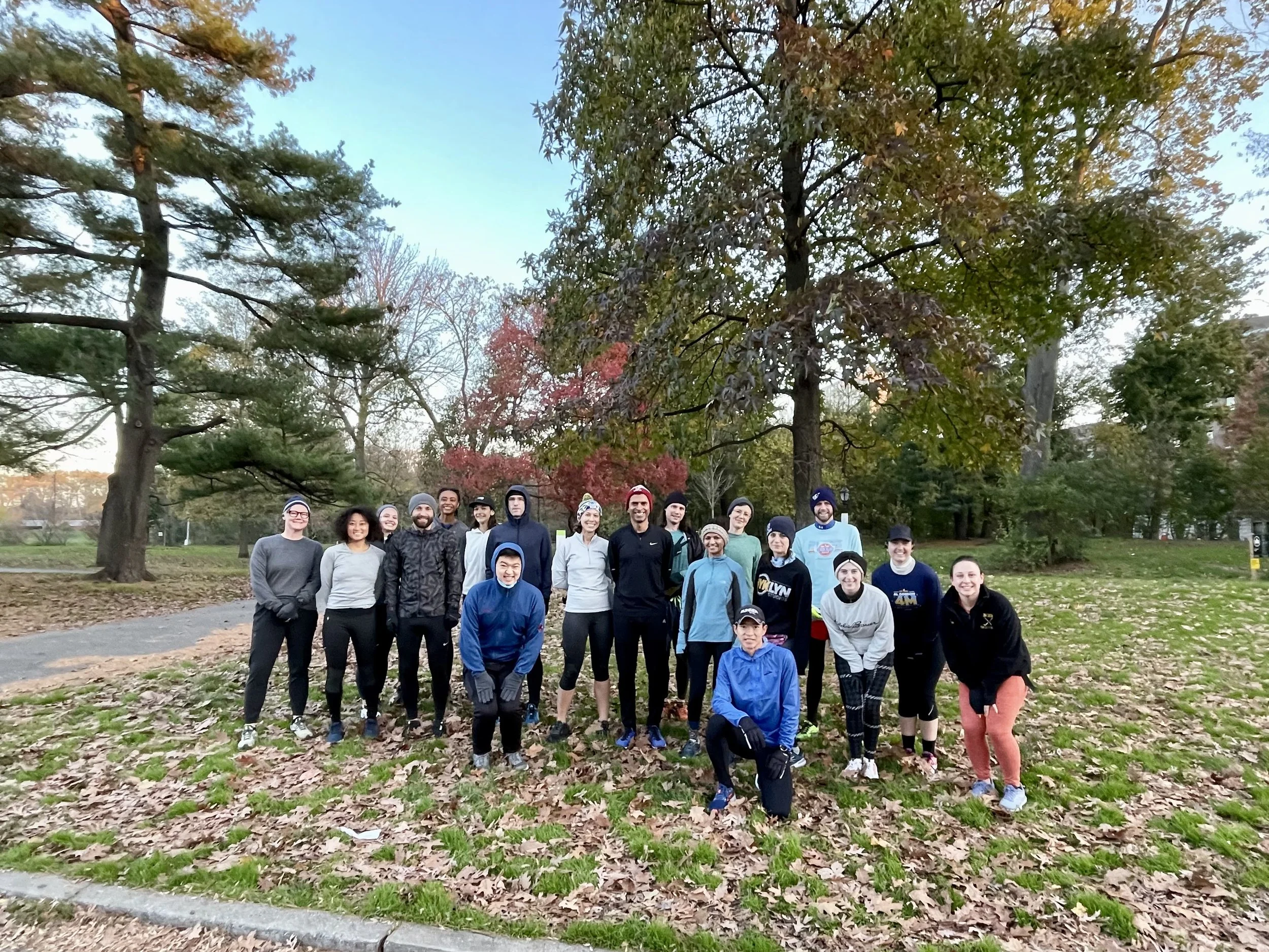 A group of about 20 bundled up runners standing and kneeling on grass strewn with brown leaves. The trees in the background have brown and red leaves or are evergreens and the sky is a bright blue.