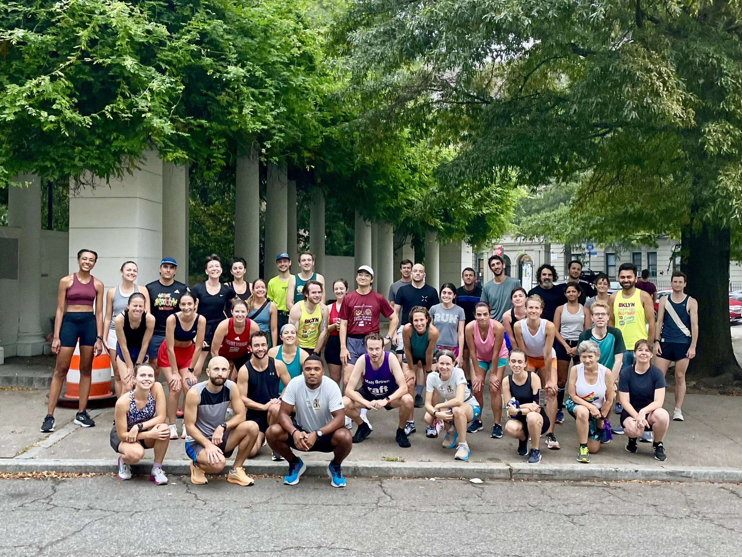 A group of about 40 runners standing next to and in front of lush green trees and vines as they stand on a concrete plaza.