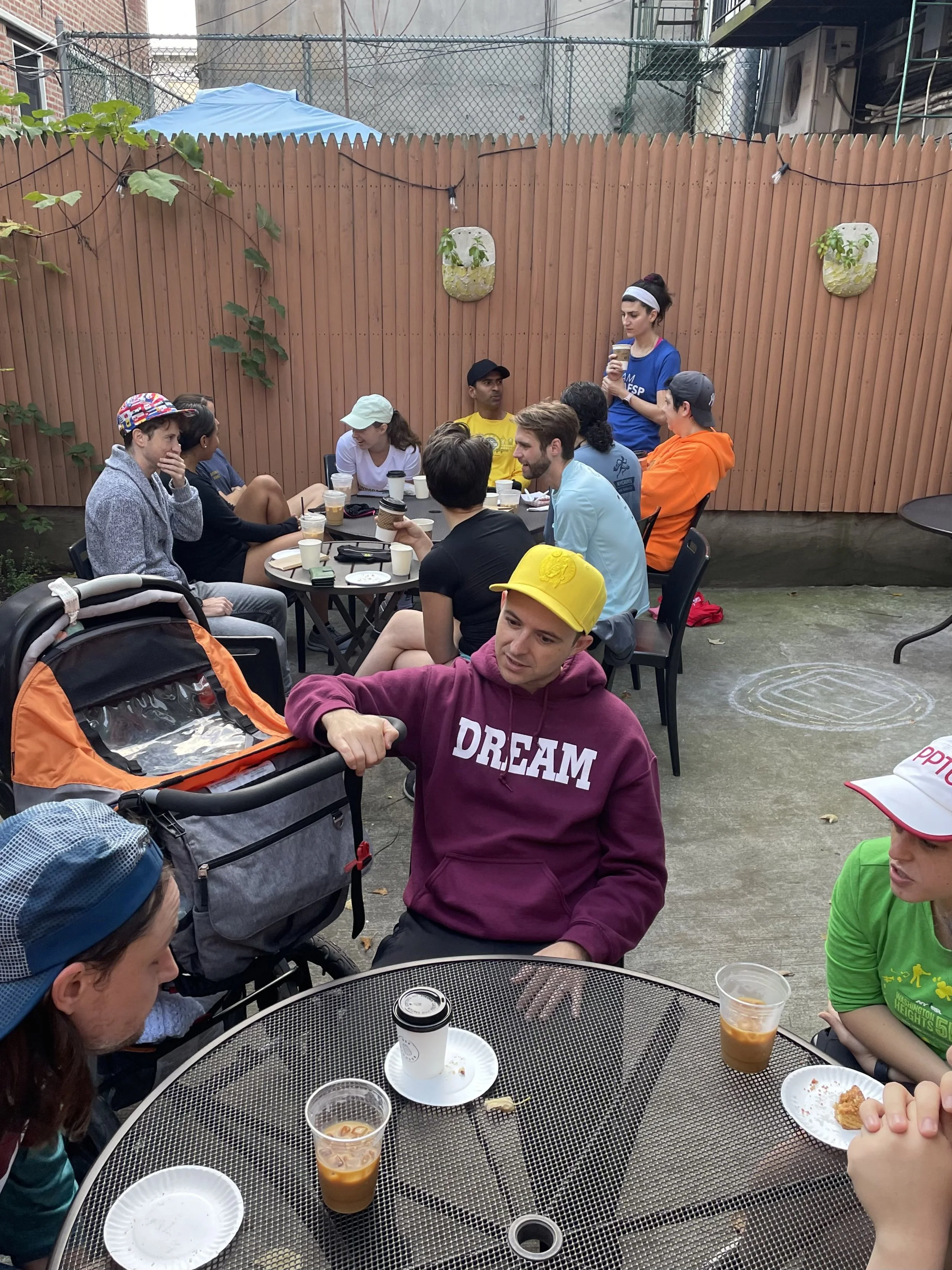 A group of about 15 runners and a baby in a stroller sitting around black metal tables outside in a wooden fenced in area with a concrete ground and pops of plants around. The tables are litered with coffee cups & pastries; the runners are conversing
