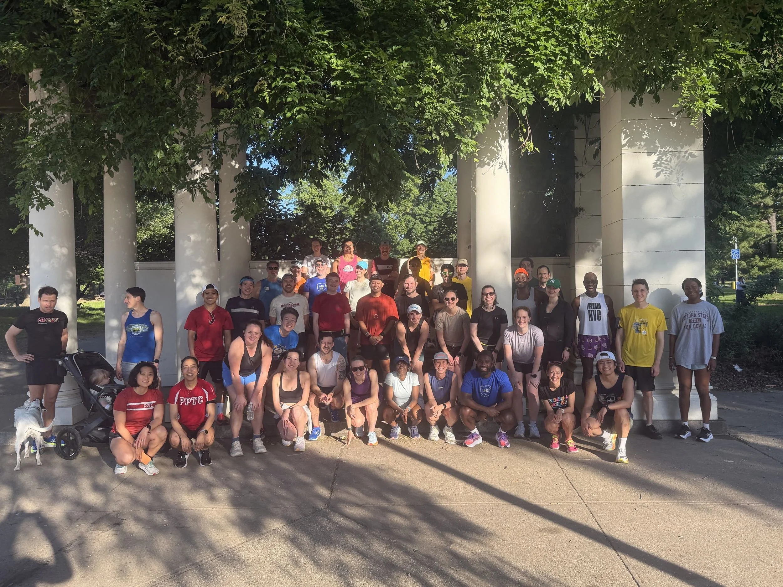 A group of about 45 runners, a baby, and a dog, underneath the pergola in dappled sunlight with lush vines cascading down from the pergola's roof.