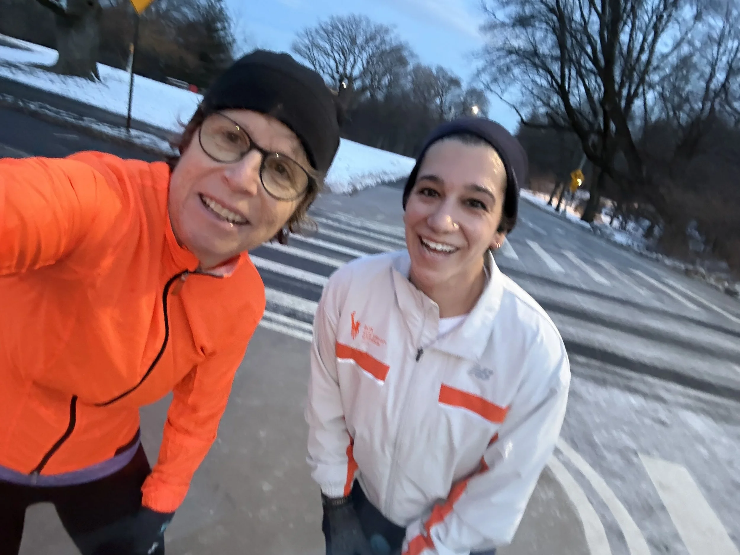 Two people in windbreaker jackets smile as they stand on the road of a snow-covered park.