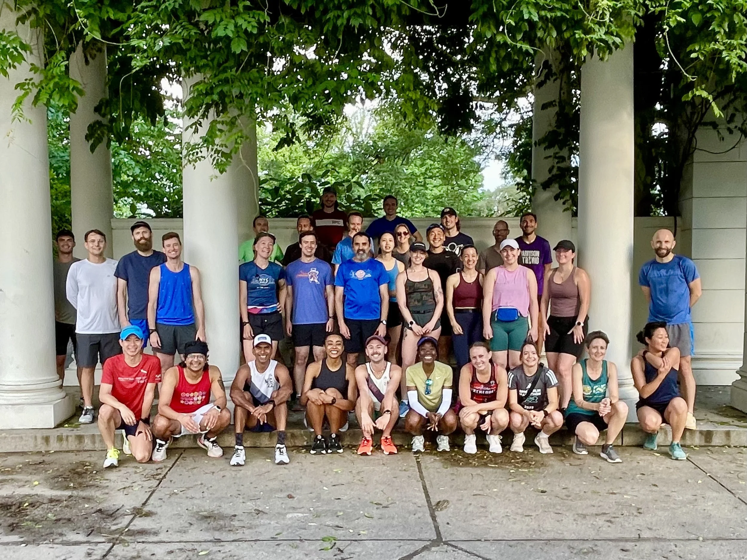 A group of about 35 runners underneath a pergola, above and behind them are vines and trees covered in deep green leaves.