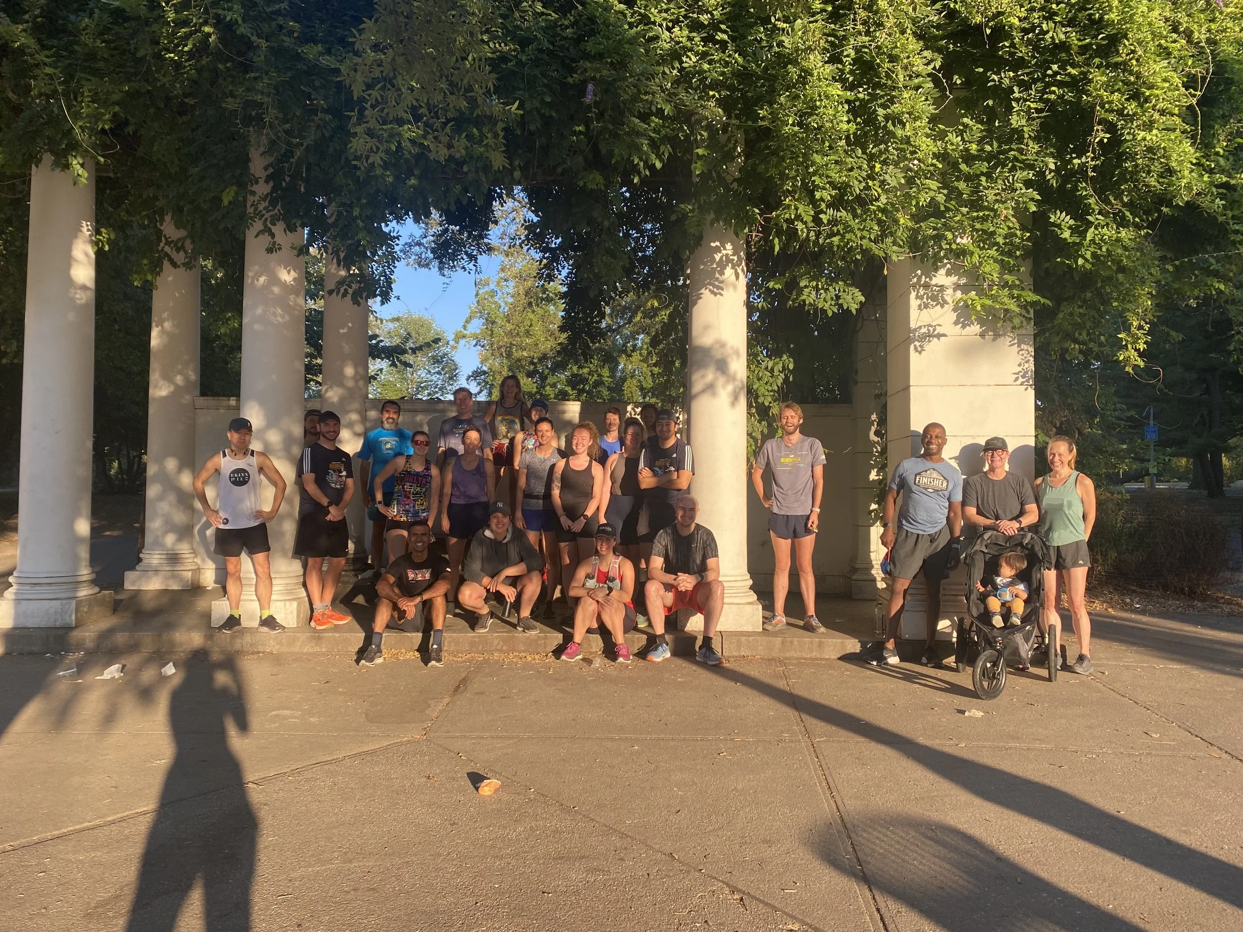 Long shadows and golden sun cast over a group of 20ish smiling runners including a baby in a stroller standing and sitting under a pergola which has an abundance of lush, leafy green vines cascading down from the roof.
