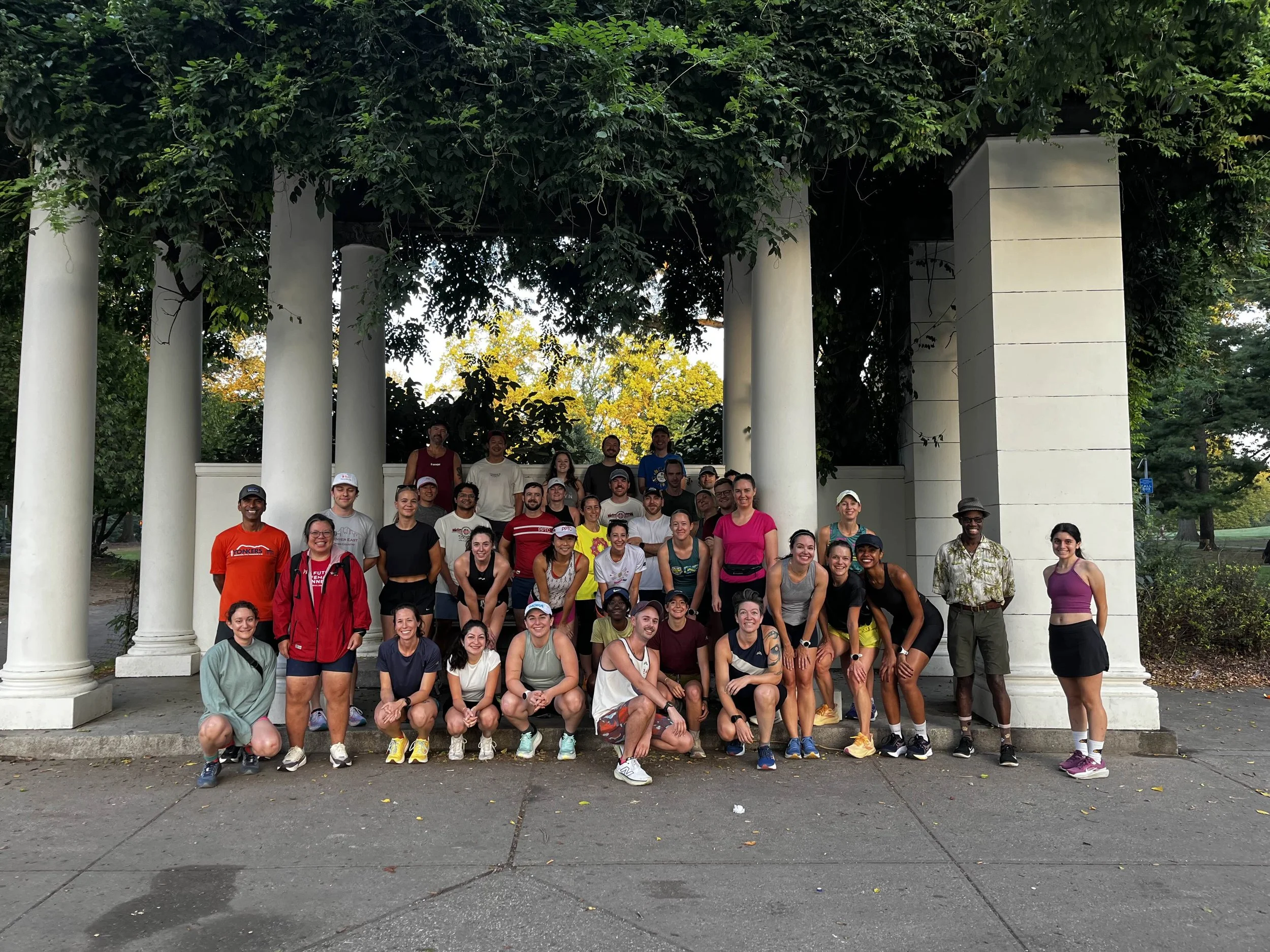 A group of about 40 runners smiling and posing underneath a pergola whose roof is a vibrant and verdant .
