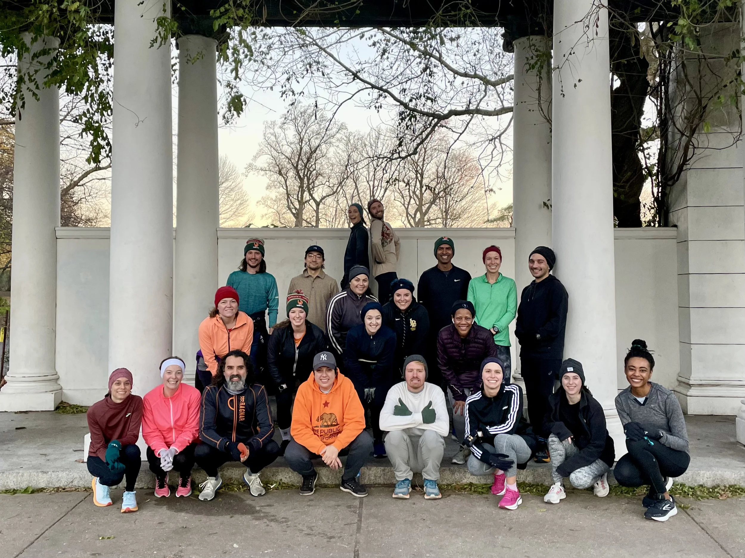 A group of about 20 runners sitting and standing under the pergola. Two in the far back are posing back to back. Behind the pergola you can see bare trees and the first traces of light.