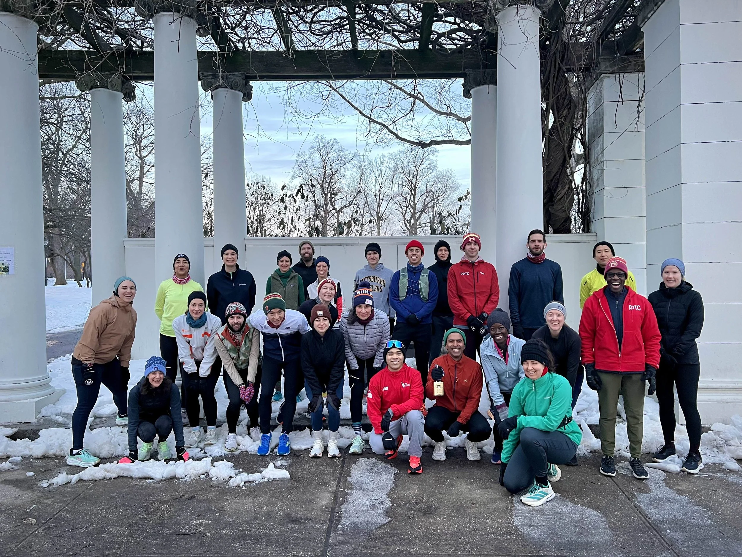 A group of 26 bundled runners gathered underneath a pergola. Snow is on the ground and one of the runners is holding a golden cowbell and smiling.
