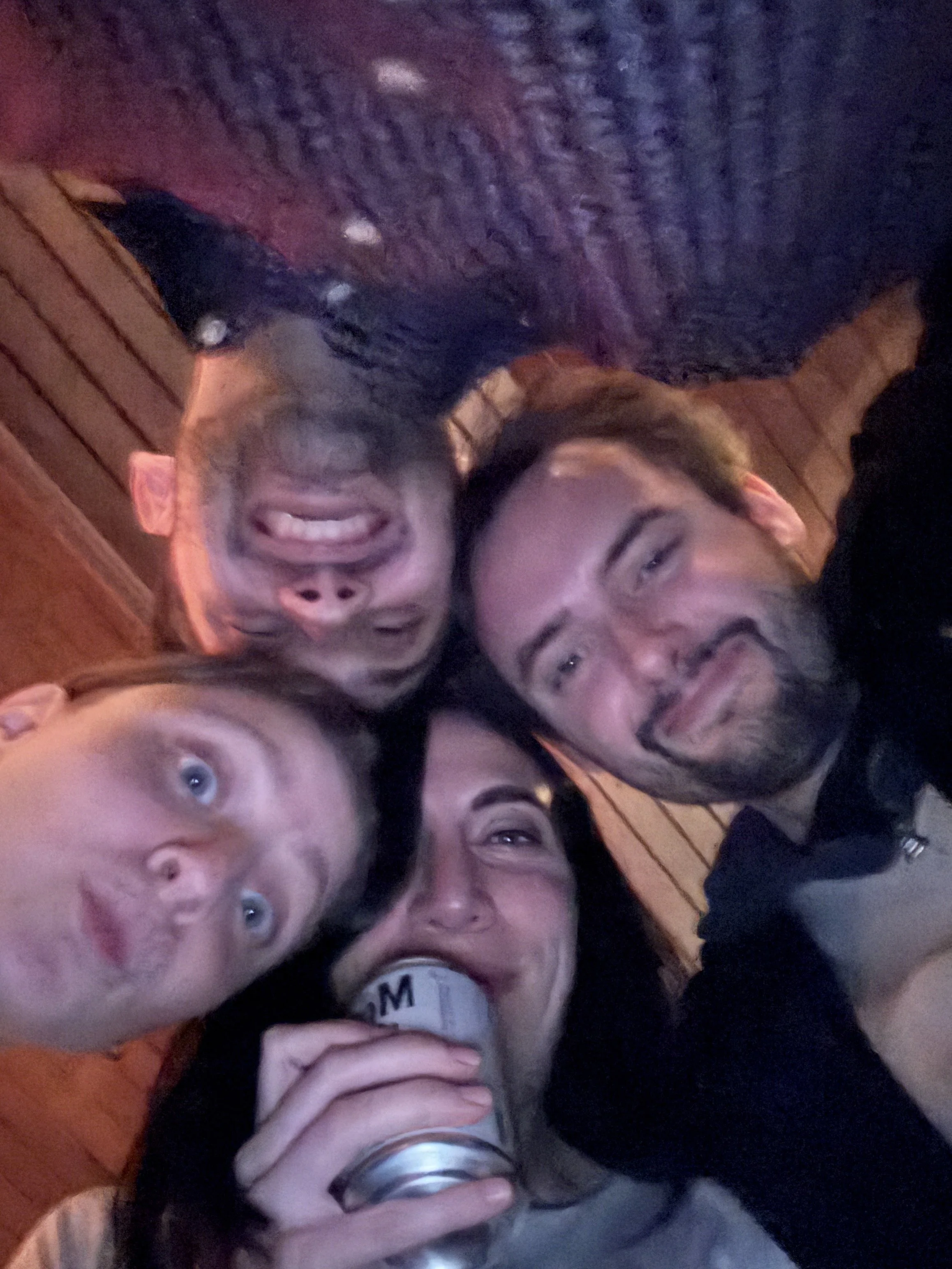 Four people touch their heads together against a wooden background as they laugh and make funny faces.