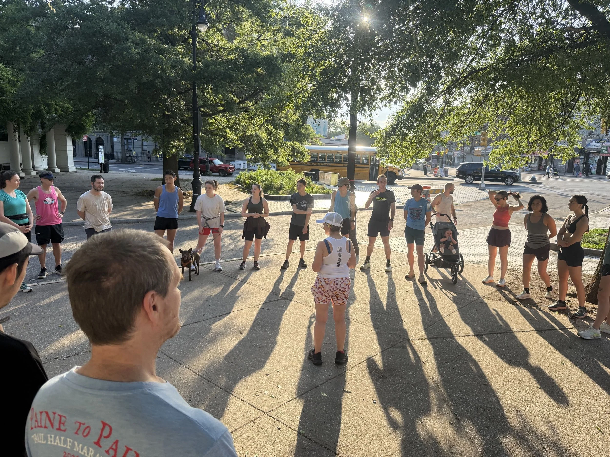 The circle up at SEC that happens before each run. In the photo you can see runners standing in a circle and their shadows spilling forward as the sun hits their backs. In the middle one runner stands and one runner has his baby in a stroller.