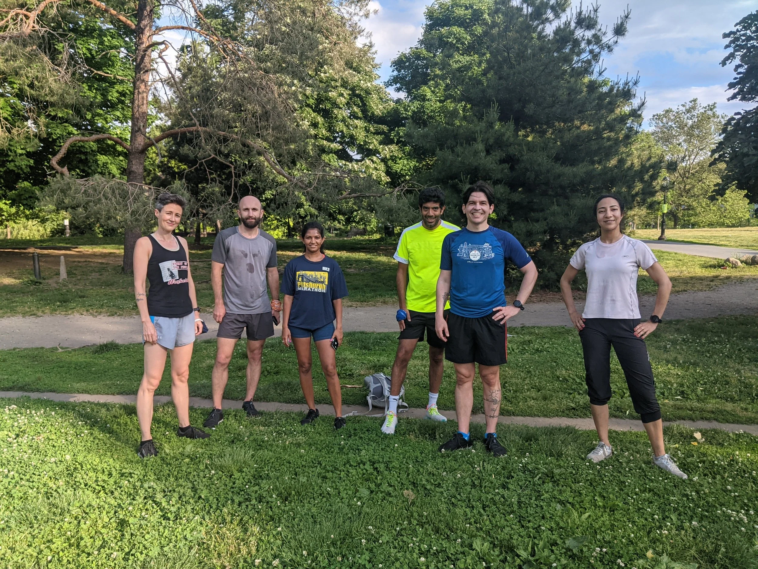 Six people standing on grass in front of a group of trees and intersecting paths. The people are smiling and wearing running clothes and standing in the shade near a patch of sun.