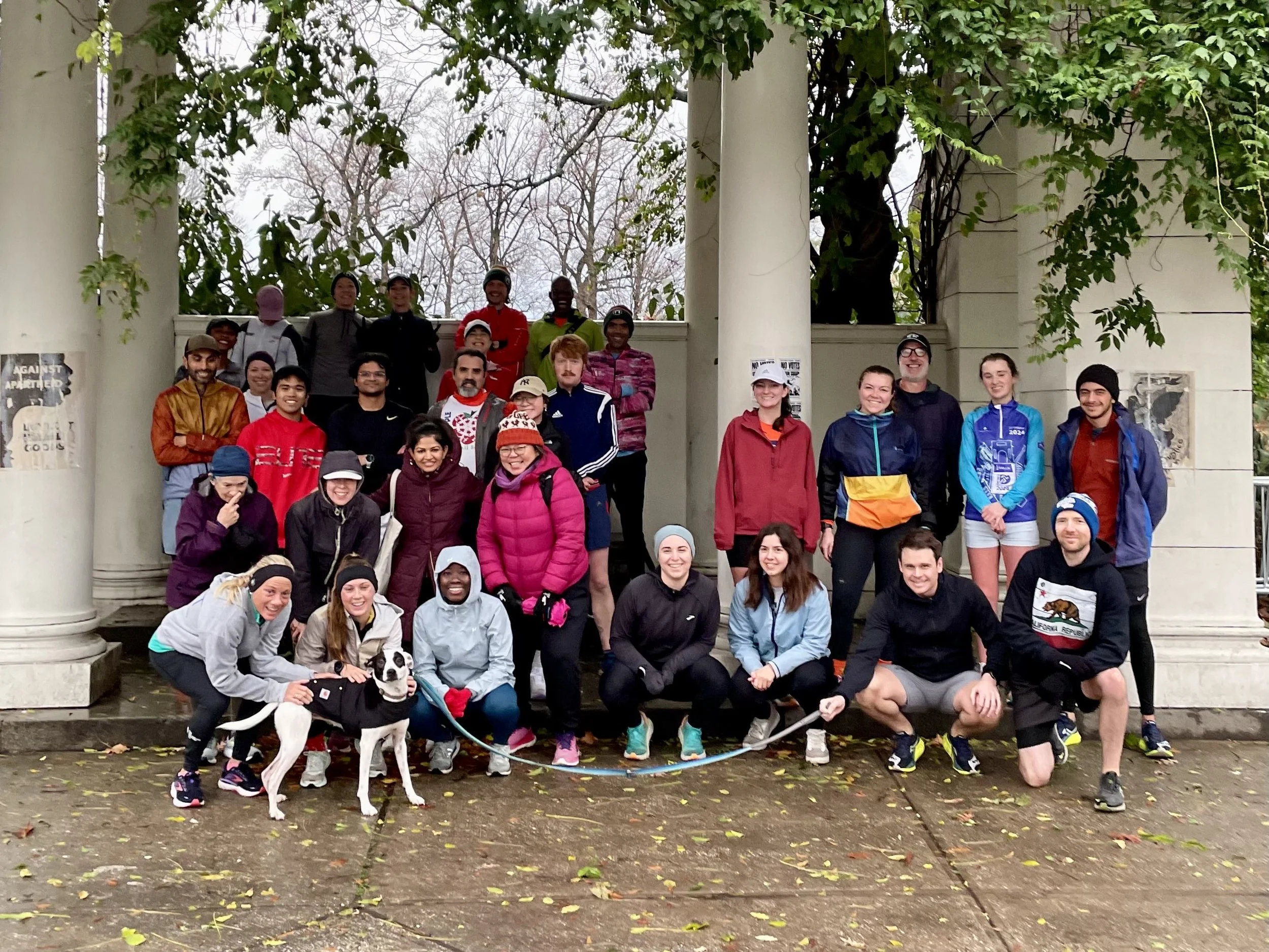 A group of about 30 runners in rain jackets, mittens, hats, and headbands are gathered underneath a pergola. Two are petting a dog on a leash that is being held by someone several people down from them.