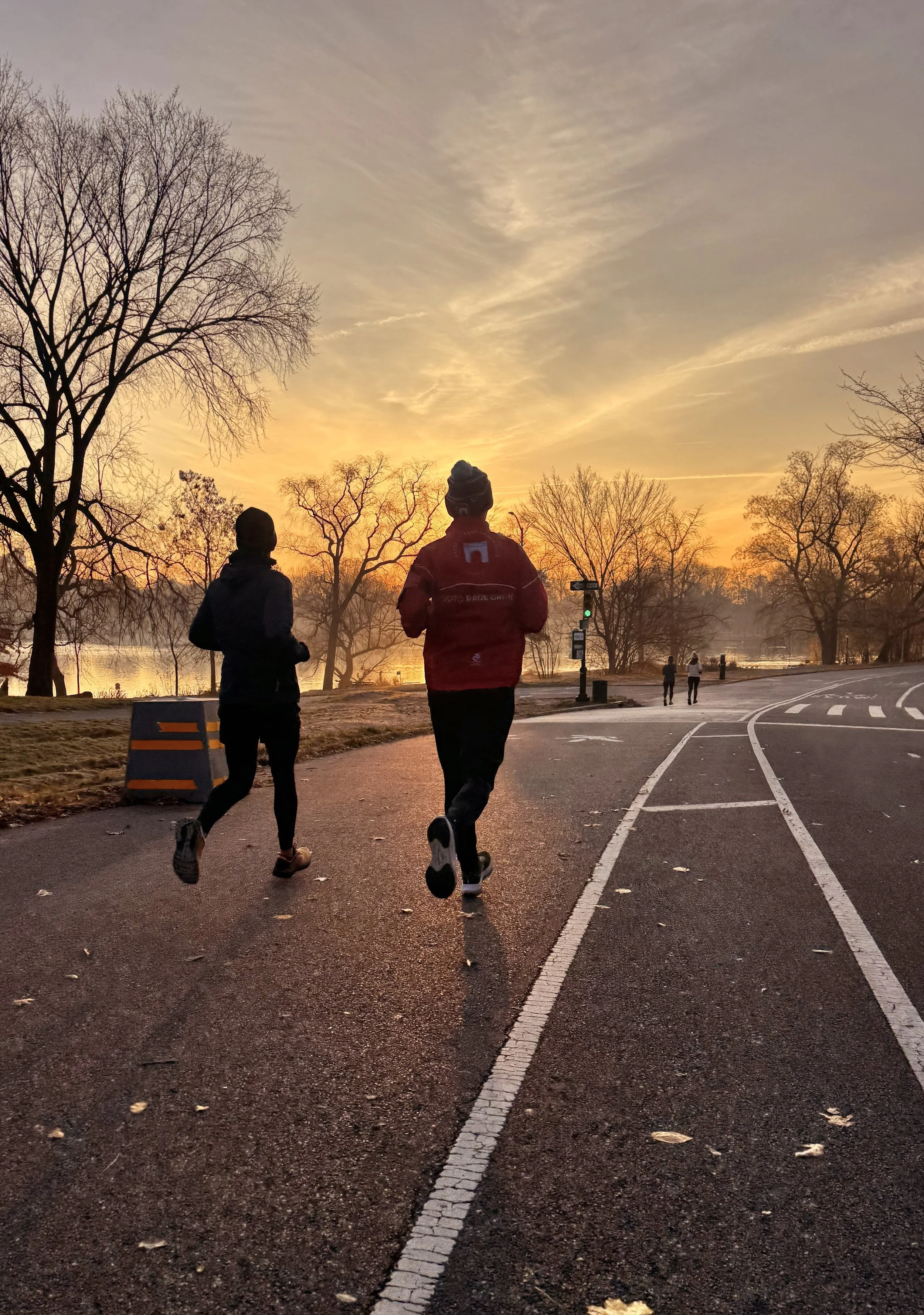Two runners bundled in winter hats and one wearing a red PPTC jacket, running on asphalt near a lake. The sky is a soft yellow and orange with streaks of wispy clouds . Ahead of them you can green traffic light and a couple other runners.