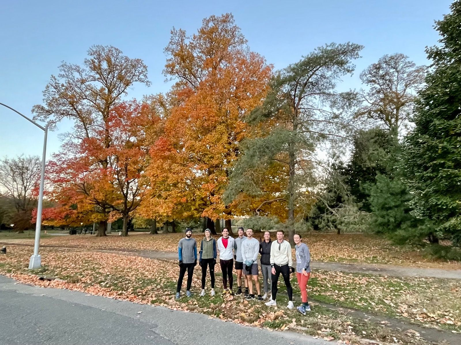 A group of eight people in running clothes stand in front of a group of evergreen and deciduous trees with varying shades of orange leaves.