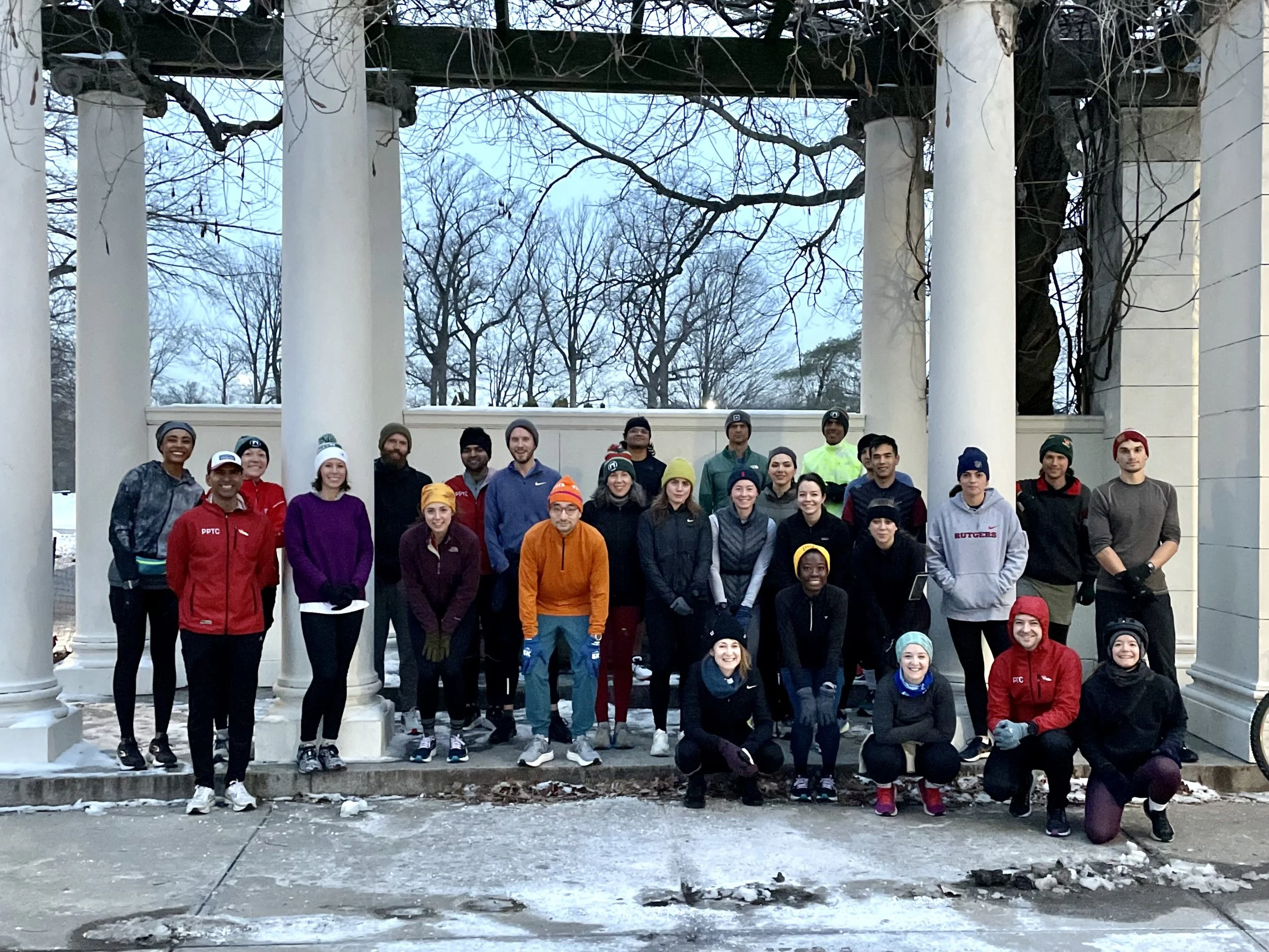 A group of nearly 30 runners bundled up underneath a pergola as salt and ice and snow lightly coat the concrete ground in white. The trees behind them and the vines above all have bare branches.