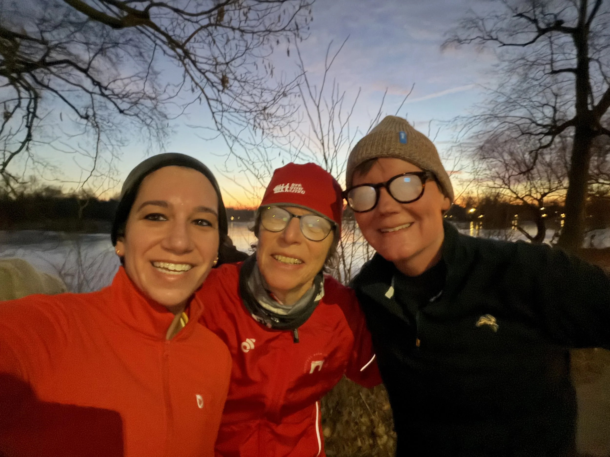 Three people stand smiling as the sun rises (or sets) over a body of water behind them. Trees with bare branches surround the water.