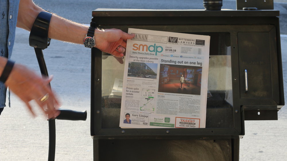 Josh holds the newspaper in front of a news stand