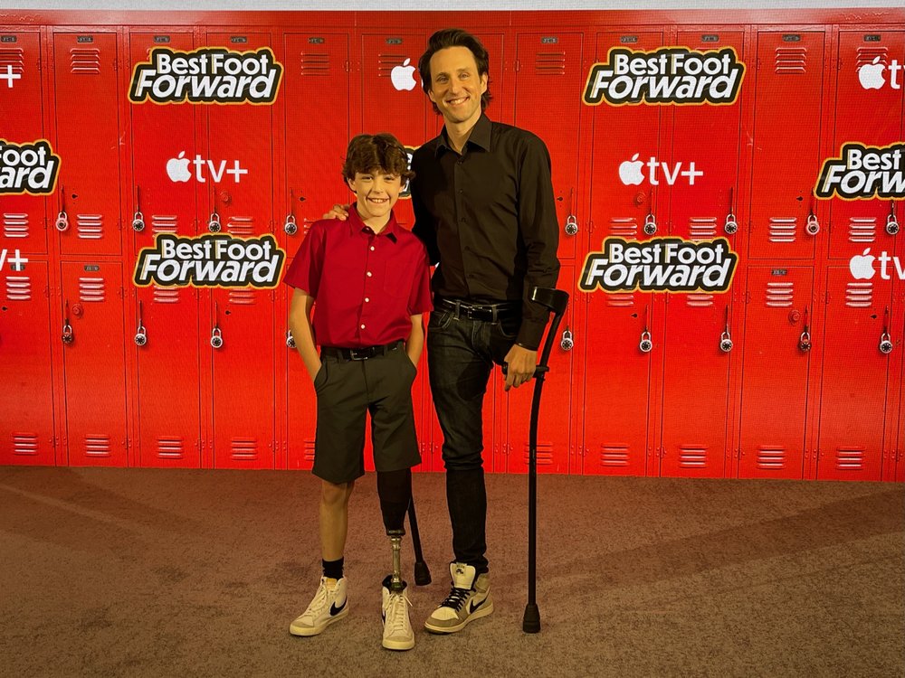 This is a photo of a young boy and a man standing next to each other in front of lockers that say "Apple TV+ and Best Foot Forward." The little boy is wearing a prosthetist on his left leg and the man is missing his left leg.