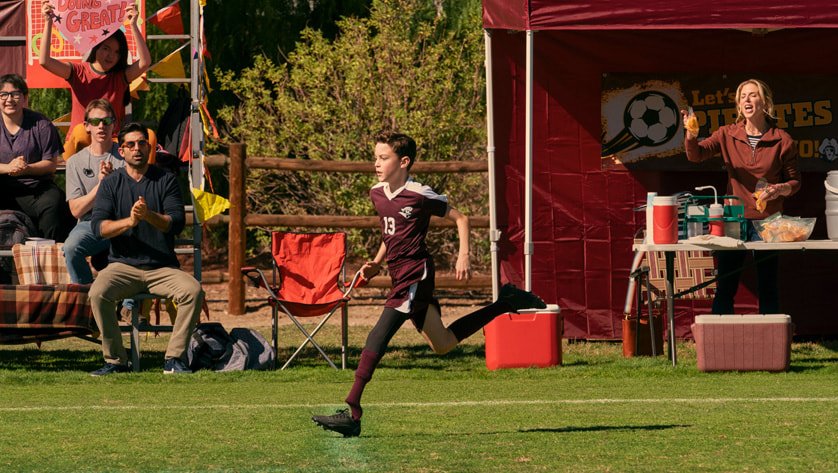 There is a young boy running on a prosthetic leg on a soccer field. There are adults sitting on bleachers cheering him on! There's also a table in background with beverages to help the athletes.