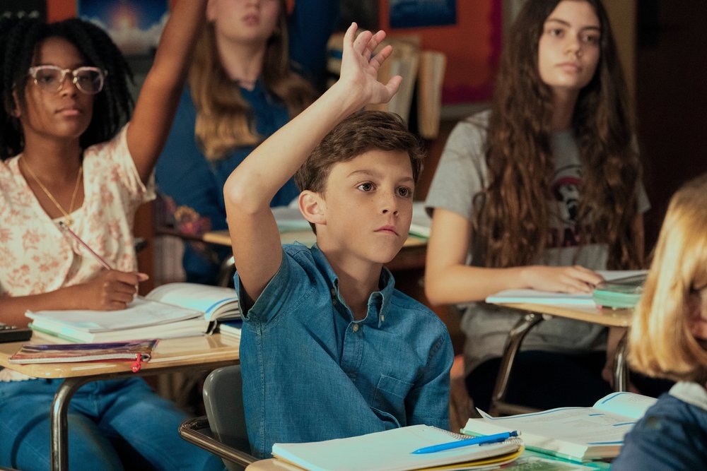 A young boy in sitting at a school desk and raising his hand. He is wearing a collared shirt. There are a few girls seated in desks around him.