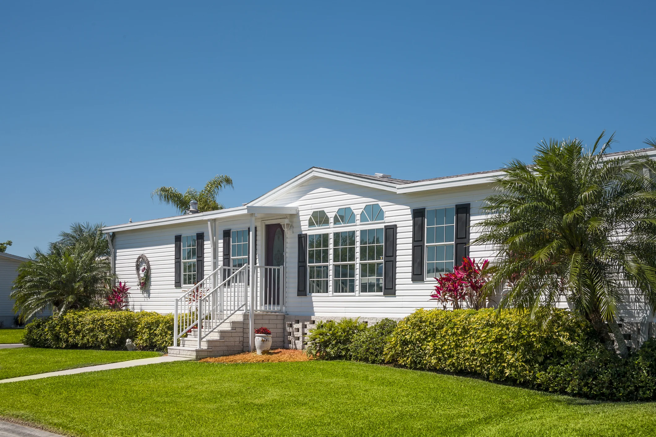 A white mobile home with a green lawn and shrubbery surrounding it