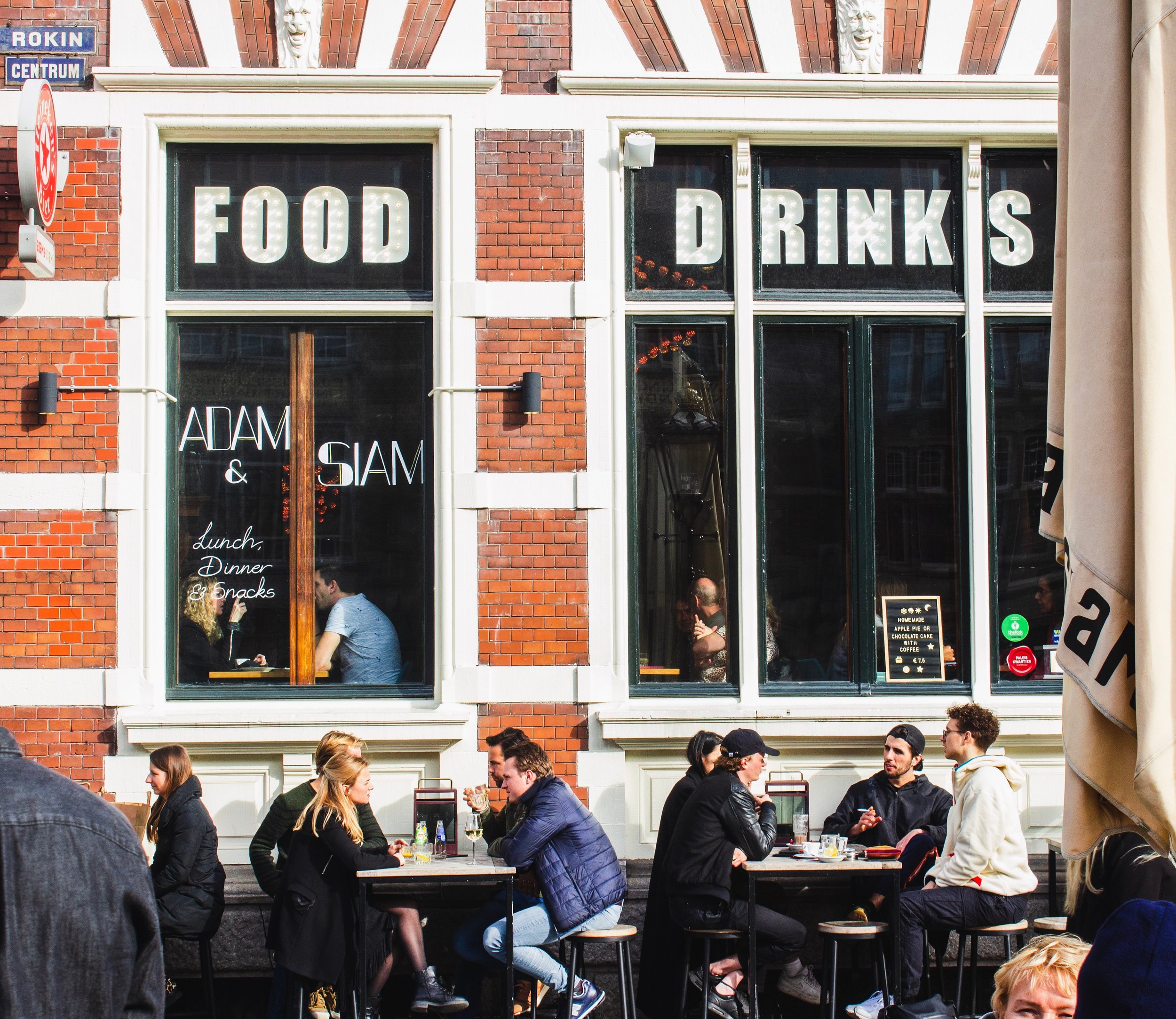  A street view of people dining at tables on the street in front of a brick building with two big windows with the words “FOOD, DRINKS” written on them  