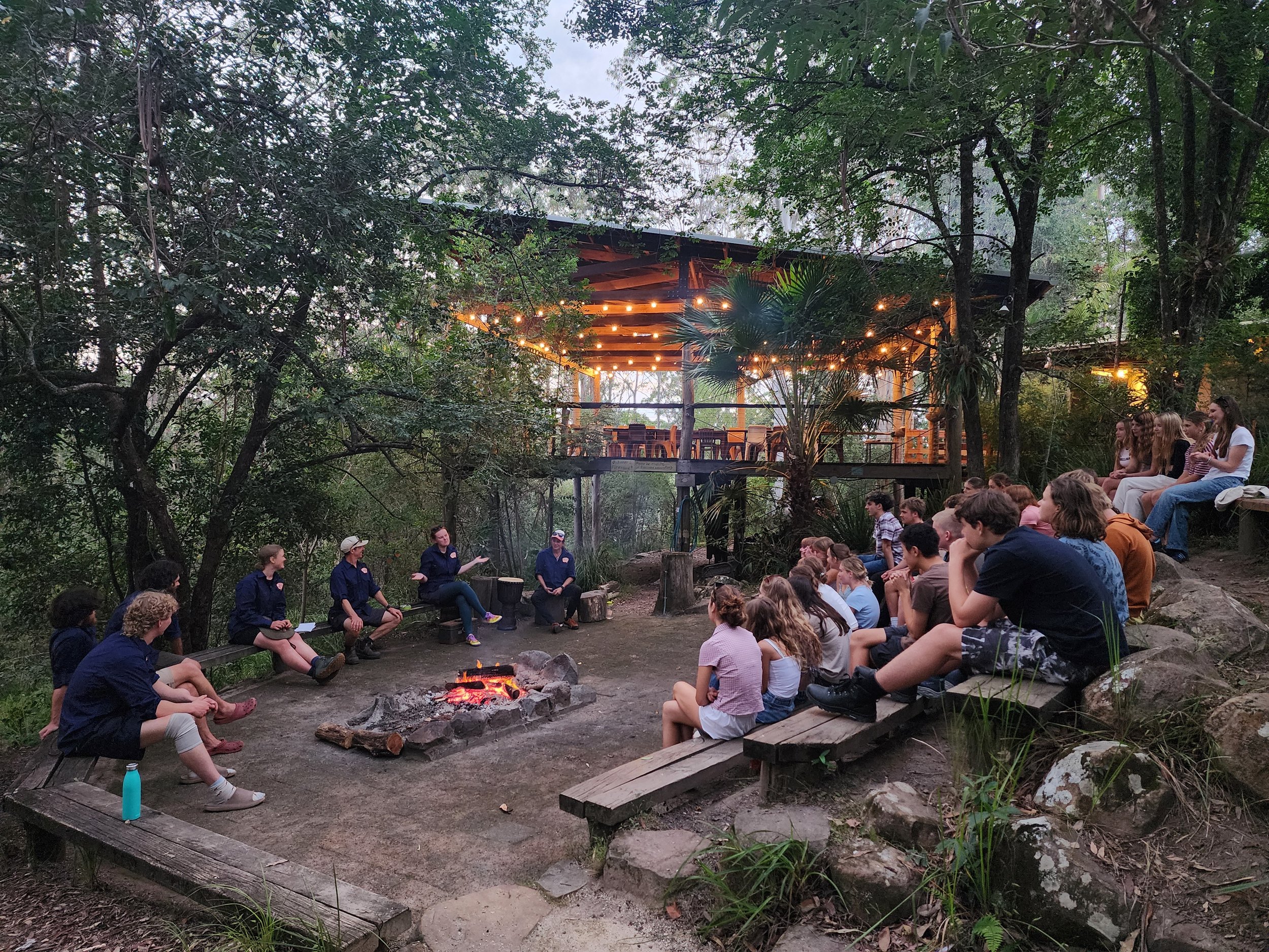 Group sitting around fire, with large deck and forest, and Wangat Lodge, Hunter Valley, Barrington, NSW