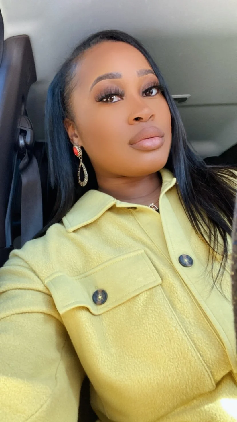 A woman with long black hair and makeup, wearing red and gold earrings and a yellow jacket, taking a selfie inside a vehicle.