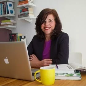 Woman sitting at a desk with a laptop, books, and a yellow mug.