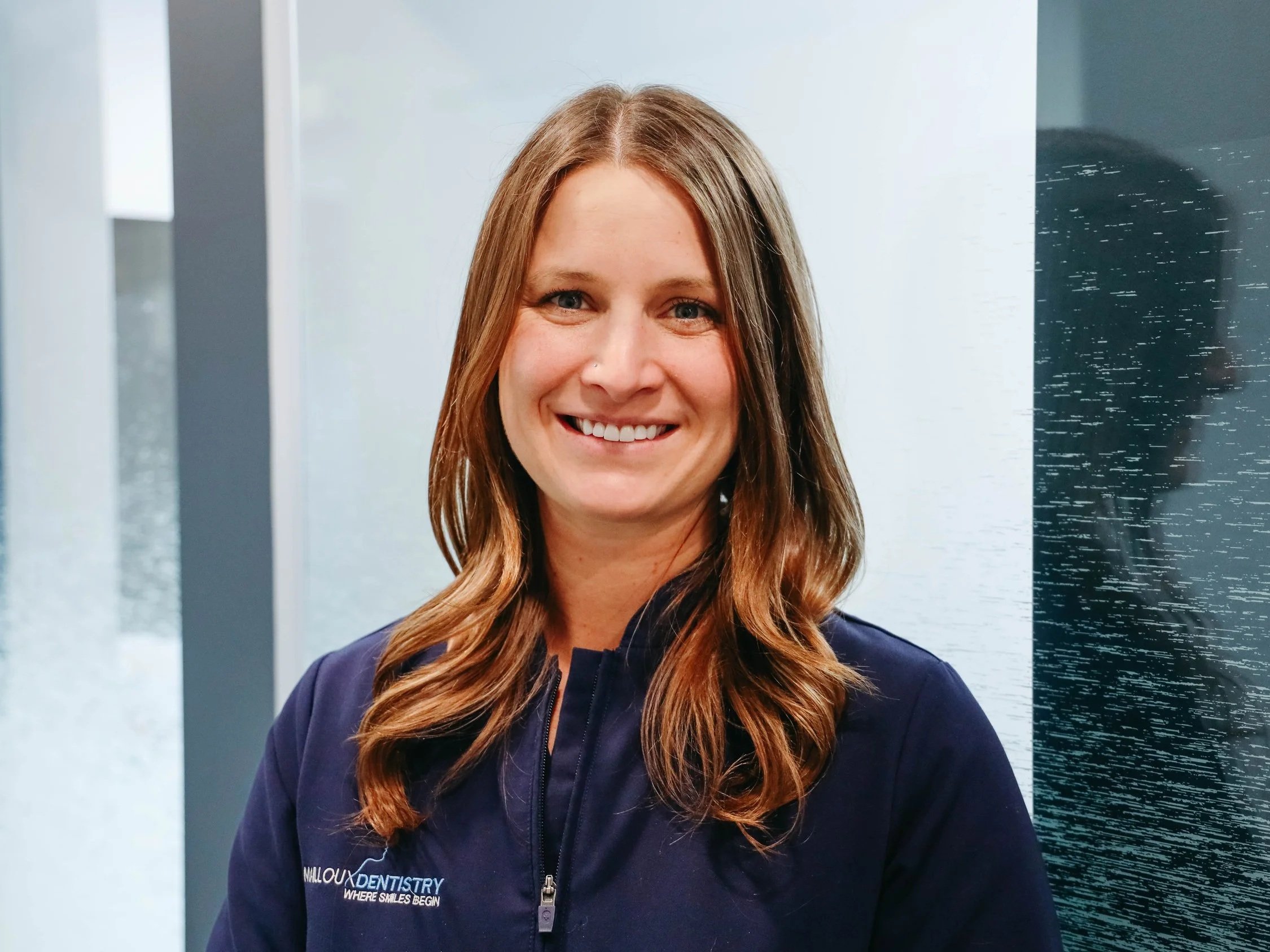 A smiling woman with straight shoulder-length blonde hair, wearing a black V-neck shirt, poses against a dark cloudy background.