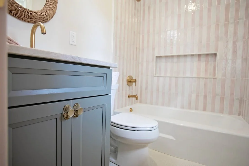 The soft blue cabinetry is accented by decorative brass hardware and paired with a white countertop, creating beautiful contrast against the blush pink and white striped wall tile surrounding the bathtub.