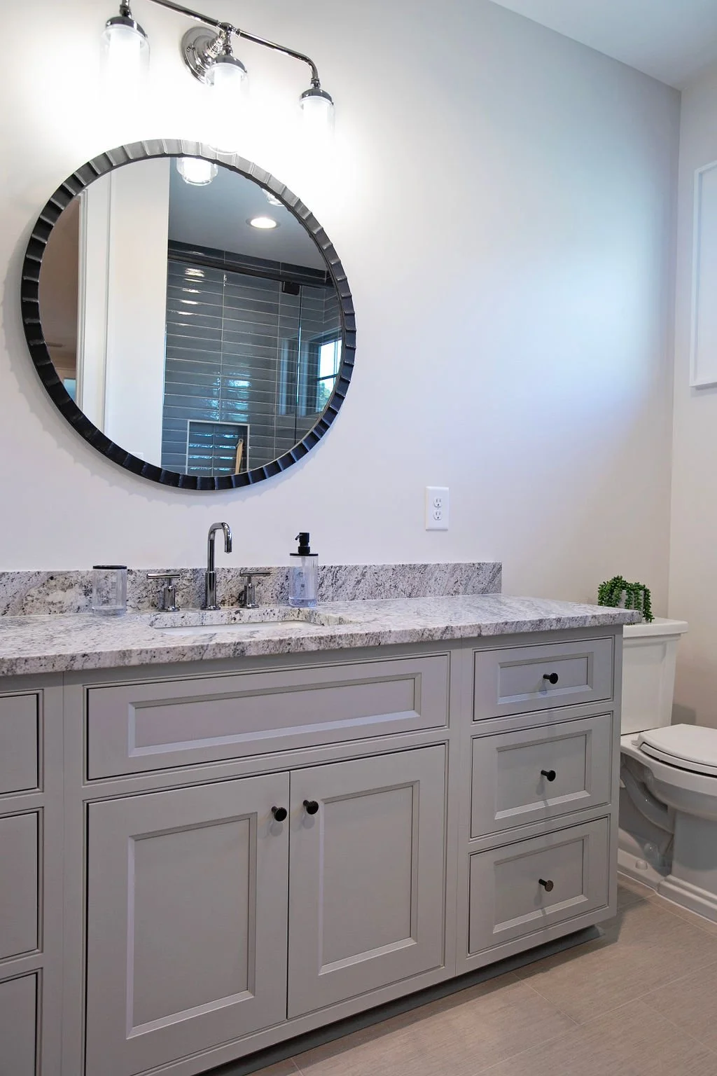 Bathroom vanity with a granite countertop, a round mirror with a black frame, and a white toilet. There is a plant on the right side of the toilet.